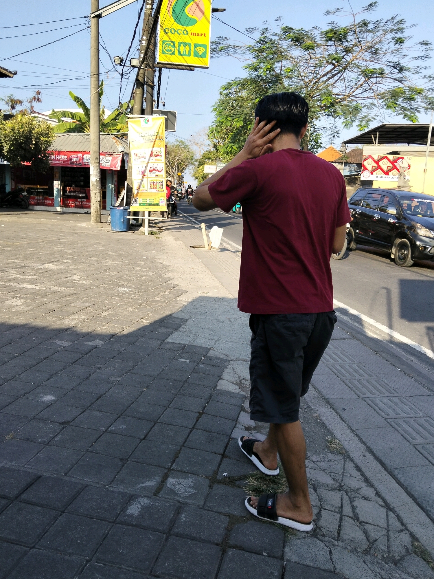 A young man walks along a tiled sidewalk in Kuta Selatan, Indonesia, with his hand behind his head. He is wearing a maroon t-shirt and black shorts. The sidewalk is made of dark gray square tiles and there is a street with a black car driving on it to his right. To his left, there are shops with awnings and advertising banners. A large tree with green leaves is visible in the background. The sky is clear and blue.