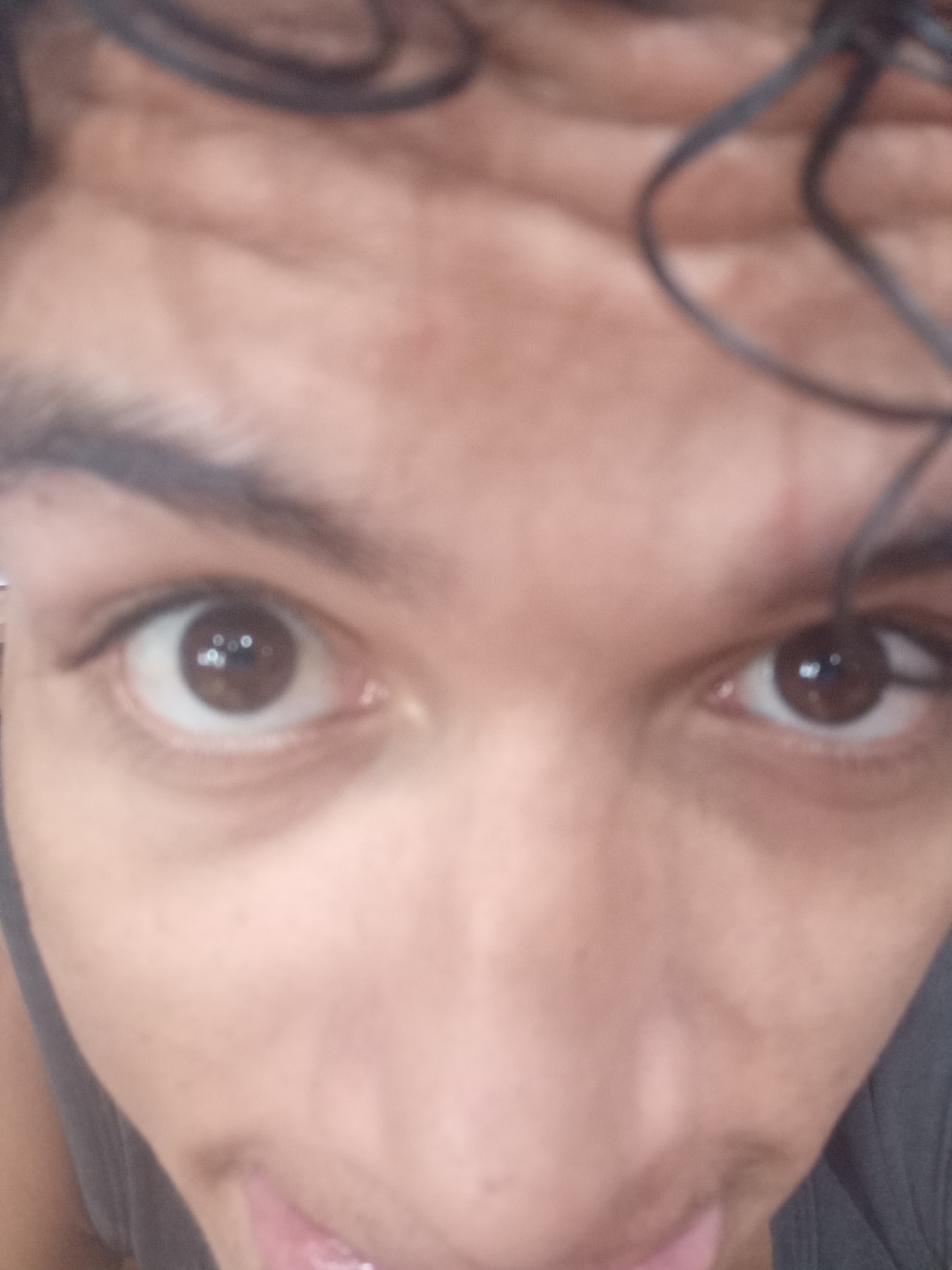 A close-up photograph focuses on a person's face, specifically the eyes, eyebrows, and the upper part of the nose and mouth. The person has brown eyes with light reflections visible in the pupils. Dark eyebrows are prominent above the eyes. The skin tone is light brown. Curly dark hair is visible at the top of the forehead and in the upper right corner of the image, suggesting the person has glasses resting on their head. The bottom of the frame shows a glimpse of the person's tongue. The image angle suggests the photo was taken from slightly above and close to the subject. The photo does not contain information to place the person in Juan Griego, Venezuela, or any other specific location.