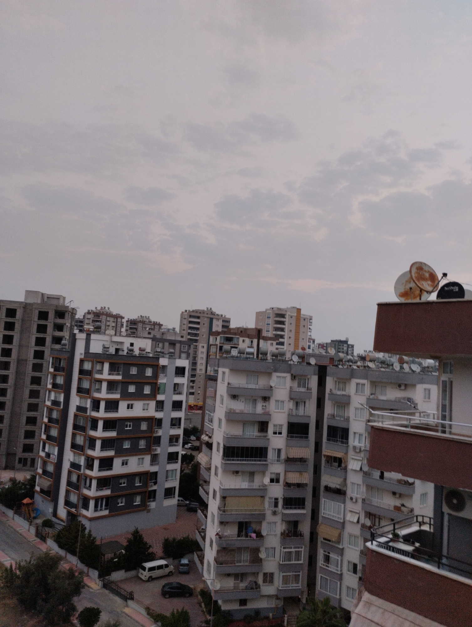 The image depicts a view of apartment buildings in the city of Mezitli, Turkey. The sky is overcast with a light gray hue, suggesting it might be dawn, dusk, or a cloudy day. Multiple residential buildings of varying heights and architectural styles are visible. Some buildings are under construction, indicated by their unfinished facades and empty window frames, while others appear occupied with visible windows, balconies, and satellite dishes on their rooftops.
In the foreground, a building with a distinctive reddish-brown facade is prominent. Below and to the left of this building, several other apartment blocks are arranged. One building in the mid-ground features a modern design with alternating bands of dark gray and white, accented by brown window frames and balconies. Further back, more buildings stretch into the distance, some with similar repeating window patterns and balconies.
At the base of one of the buildings, a paved area serves as parking. A white van and a dark-colored car are parked. A narrow street or road is visible along the edge of the scene, with some trees and greenery scattered around the buildings. The overall scene conveys a dense urban residential environment.