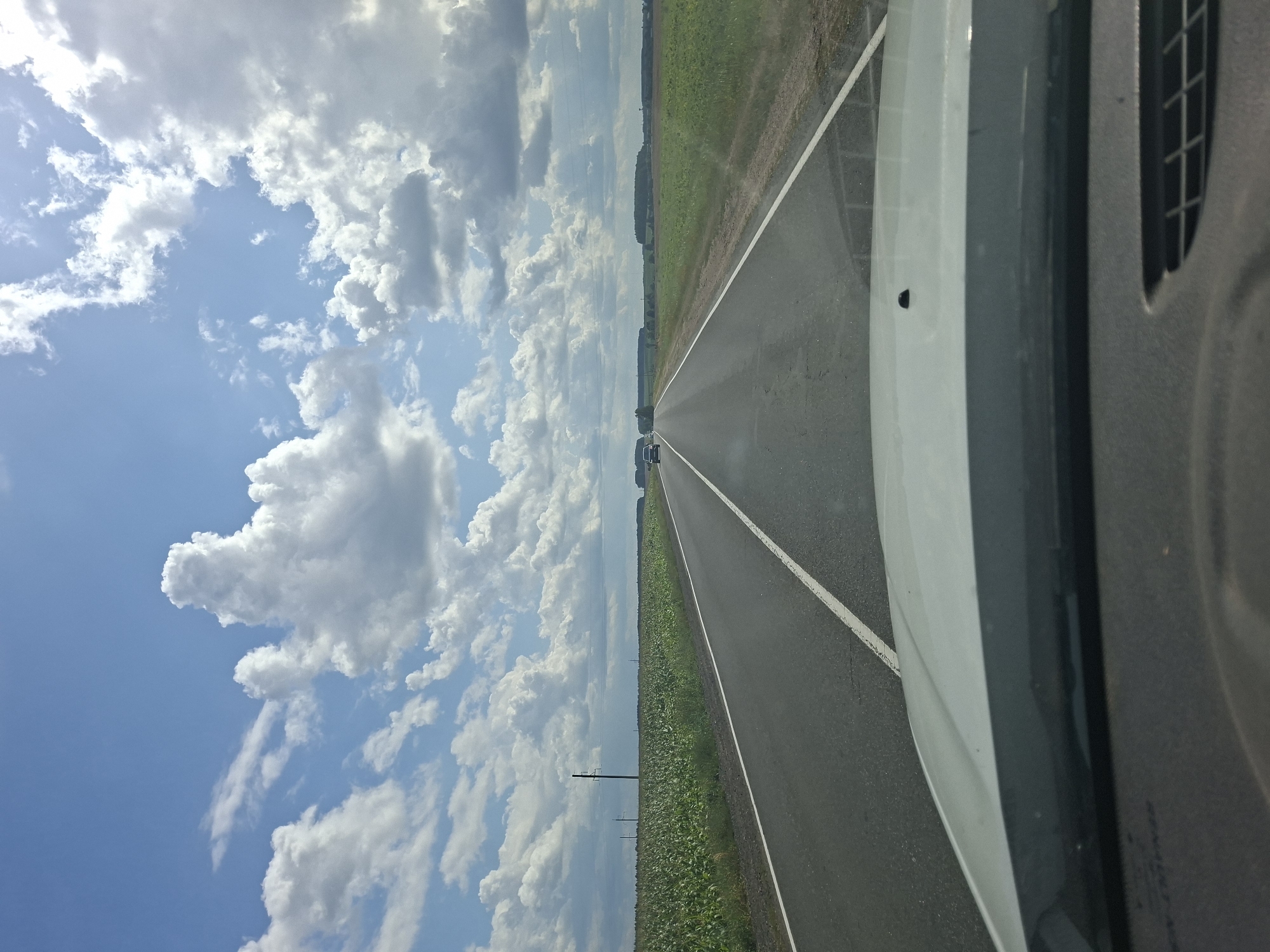 This is a first-person perspective, medium shot, outdoor photograph taken from inside a vehicle driving on a paved road. The road is asphalt with a white solid line on the right side and a dashed white line on the left side, indicating two lanes of traffic. The road stretches into the distance, curving slightly to the right.
On the left side of the road, a vast field of green crops, likely corn, extends to the horizon. Scattered throughout the field are utility poles, one prominent dark pole with crossbars stands out. Overhead power lines run parallel to the road.
On the right side of the road, another field of green crops or grass is visible, also extending to the horizon. In the far distance, a line of trees marks the edge of the landscape.
The sky is a vibrant blue with numerous fluffy white clouds, some of which are darker and appear to be approaching rain. The sun is not directly visible but its presence is indicated by the bright illumination of the clouds and the overall brightness of the scene.
Two dark-colored vehicles are visible in the distance on the road, traveling in the same direction as the camera. The foreground of the image shows the white hood and windshield of the vehicle from which the photograph is taken.
The scene is consistent with a rural setting in Belarus, possibly the Старашклоўскі сельскі Савет region, characterized by agricultural fields and open roads. The lighting suggests it is daytime, likely late morning or early afternoon. There are no people visible in the frame.