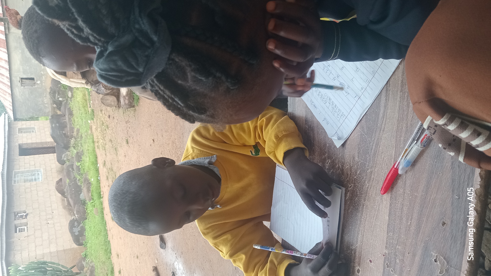 In Ribina East, Nigeria, two young children are engaged in learning activities outdoors. A boy, wearing a yellow sweatshirt, sits on the left, diligently writing in a lined notebook with a pencil. To his right, another child, partially visible and facing away, is also writing on a sheet of paper containing numbers. The setting appears to be a rural or semi-rural area, with a dirt ground and a backdrop of simple brick buildings and greenery. Two pens, one red and one blue, rest on the wooden table in the foreground. The overall scene depicts an educational moment taking place in a modest outdoor environment.