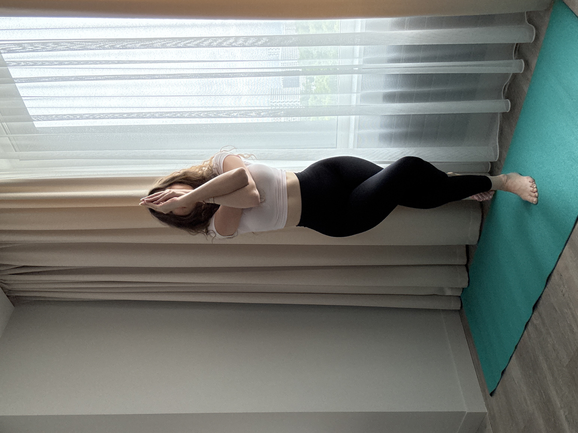 A woman with long, curly brown hair performs a yoga pose called Garudasana, or Eagle Pose, on a teal yoga mat in front of sheer white curtains and tan drapes. She is wearing a light pink cropped t-shirt and black leggings. Her right leg is crossed over her left leg, and her arms are also crossed, with her hands pressed together in front of her face. The yoga mat is positioned on a light-colored wooden floor. In the background, through the curtains, a cityscape with buildings and trees is visible. The lighting suggests it is daytime, with natural light streaming in from the window.