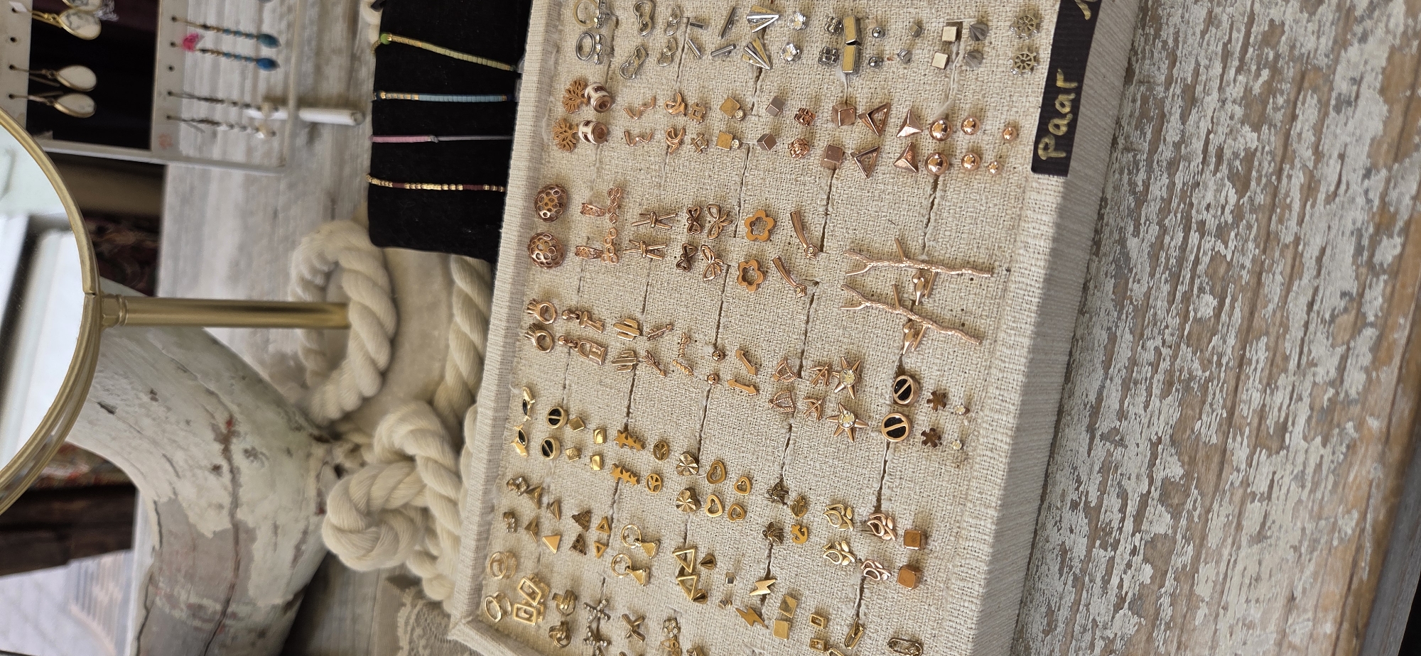 The image shows a display of various earrings on a textured fabric surface, likely in a store. The earrings are gold, rose gold, and silver, with designs ranging from geometric shapes to nature-inspired motifs like cacti and branches. Some earrings appear to be studs, while others are dangles. In the background, a rope and part of a mirror stand are visible. A black label with the word "Paar" is attached to the bottom right of the display. The display is placed on a weathered wooden surface. The scene appears to be a retail environment, possibly a jewelry store or a market stall. There is no information in the image to suggest a specific location in Karlsruhe, Germany.