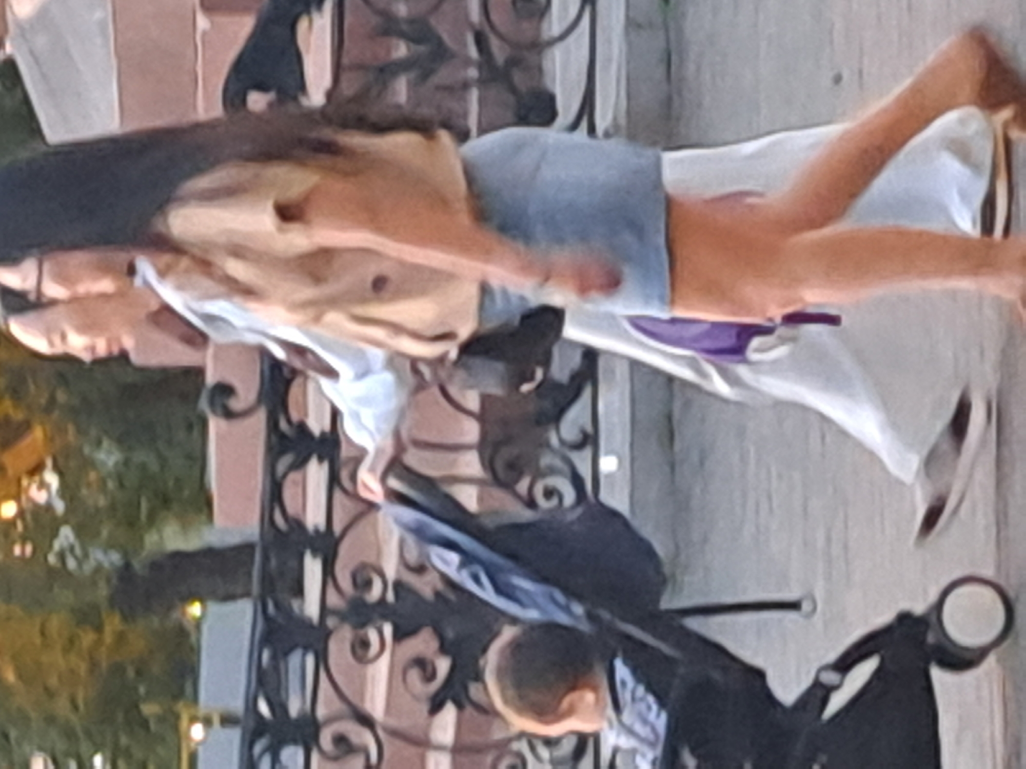 Two women and a baby are walking down a paved sidewalk in what appears to be a city. The woman in the foreground, on the right, is wearing a light brown t-shirt and a light blue denim skirt. She has long, dark hair and is wearing glasses. The woman behind her, on the left, is wearing a white shirt and dark pants. She has long, dark hair. The baby is in a stroller, and appears to be looking to the left. The background of the image shows a black metal fence and some brickwork. There are also some trees and lights in the background. The scene suggests a casual outing in a public space.