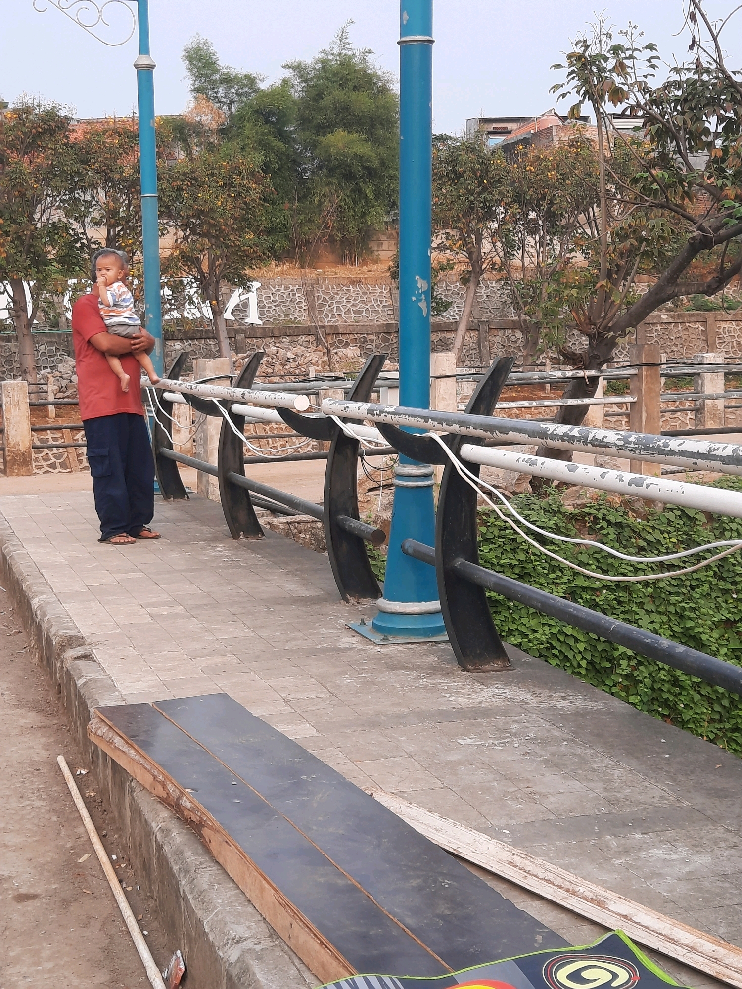 In East Jakarta, Indonesia, a person stands on a paved walkway holding a child. The person, wearing a red shirt and dark blue pants, cradles the child who is dressed in a striped shirt and gray shorts. A blue lamppost is positioned behind them, and a black metal railing with white wires runs along the walkway. Beyond the railing, a dense green hedge is visible, and in the background, trees with scattered yellow fruits and a stone wall can be seen. The walkway is bordered by a curb, on which two large wooden planks with black surfaces are placed. The ground is a mix of dirt and paving stones.