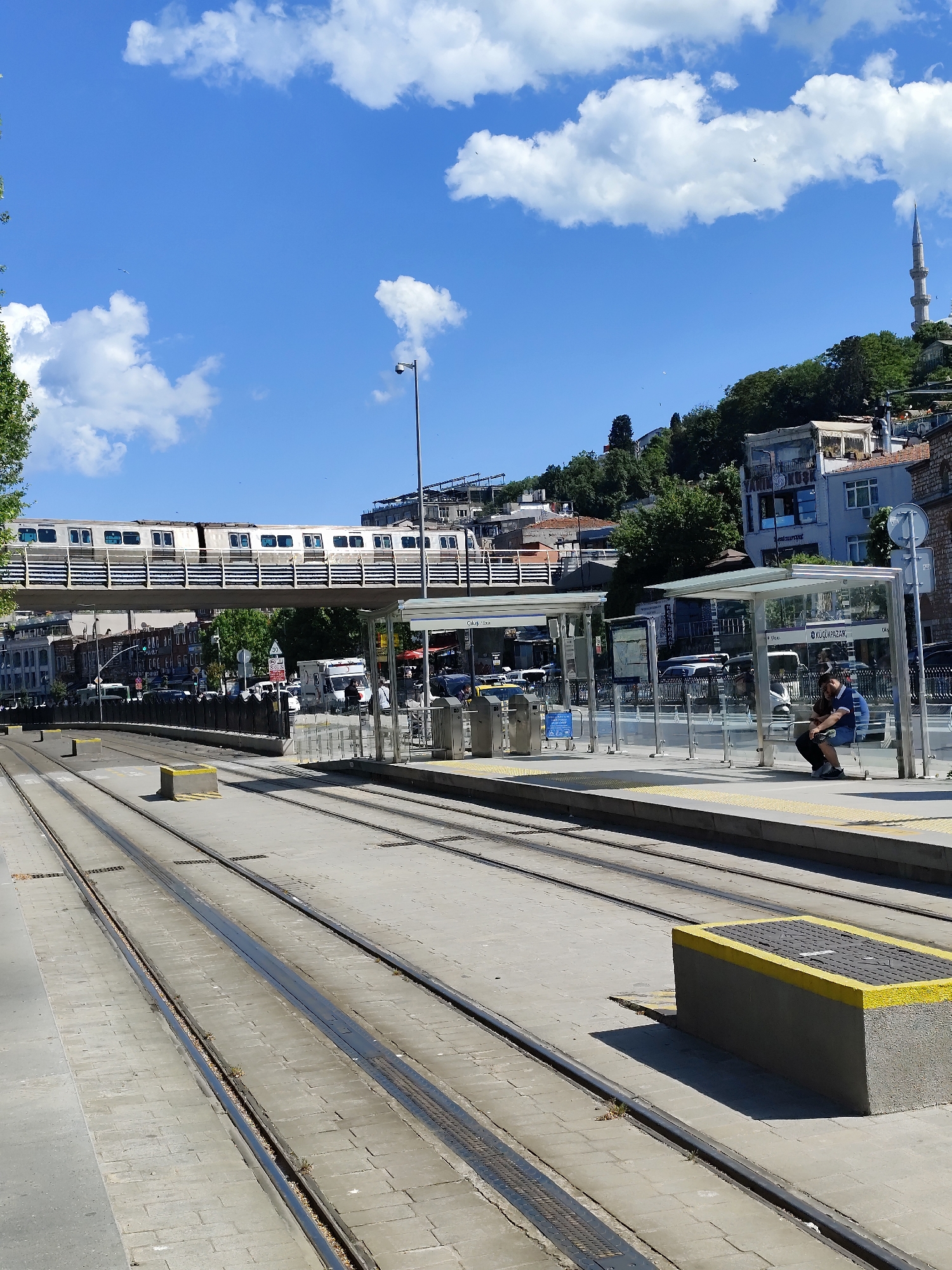 The image depicts a light rail station in what appears to be an urban setting. A person is seated on a bench under a glass shelter at the station, using a mobile phone. Tracks run along the ground, and a train passes overhead on an elevated bridge. The sky is blue with scattered clouds. Buildings and trees are visible on a hillside in the background, with a minaret visible on the far right.