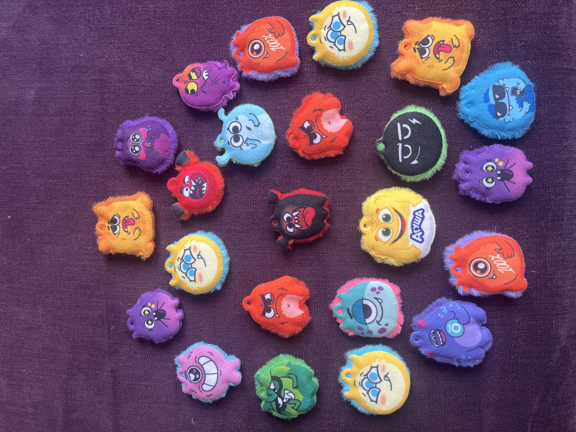 The image shows a collection of approximately 20 small, plush toys with cartoonish monster-like faces. The toys are of various colors and designs, each with a distinctive expression or feature. They are scattered on a purple, textured surface. One toy has the word "АГУША" printed on it. The image does not provide location-specific information to confirm it was taken in Krasnodar, Russia.