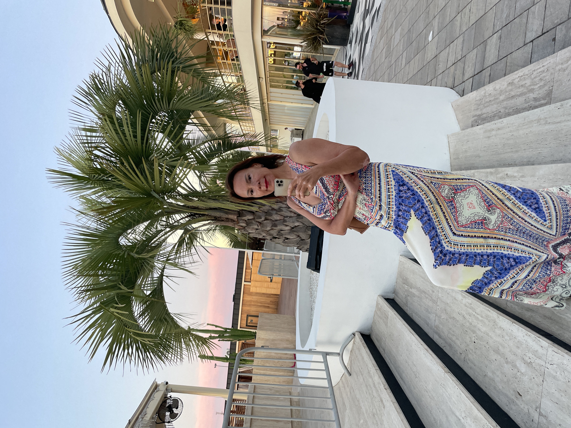 A woman with dark hair and red lipstick is taking a selfie on a smartphone. She is wearing a long, patterned dress with blue, white, and red designs. She is standing on a set of outdoor stairs with light-colored stone treads.
Behind her, a large palm tree with green fronds is prominent. Beyond the palm tree, there is a building with windows and balconies. In the background, two men in black shirts are visible, one of whom appears to be waving. The scene suggests an outdoor public space, possibly a promenade or a resort area. The sky in the background has a soft pink and blue hue, indicating it is either sunrise or sunset.
The overall impression is a casual, relaxed moment captured during an evening stroll. The location cues, such as the palm tree and the architecture, combined with the provided location context of Sochi, Russia, suggest a warm-weather setting. The woman's expression is pleasant and engaged with the camera. There is no visible text in the image.
