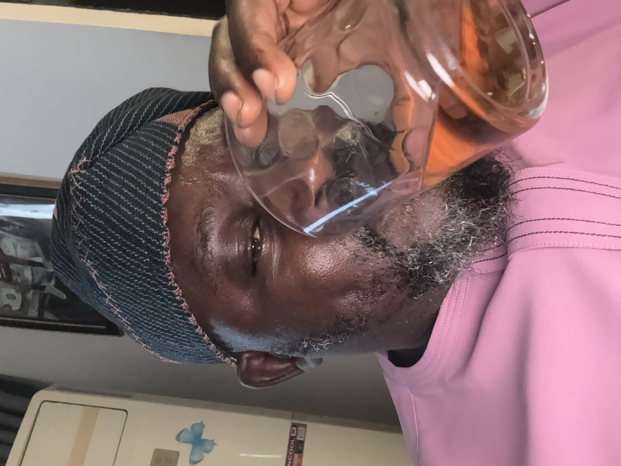 A close-up, rotated view of an adult Black male drinking from a clear glass. The glass contains a tan-colored liquid. The man wears a dark blue and reddish-brown hat and a pink shirt with a pocket. His skin is dark brown and slightly reflective, and he has a grey beard and mustache. A white appliance with a blue butterfly decoration is visible in the background. A frame is visible behind the man's hat. The location is Igboefon, Nigeria.