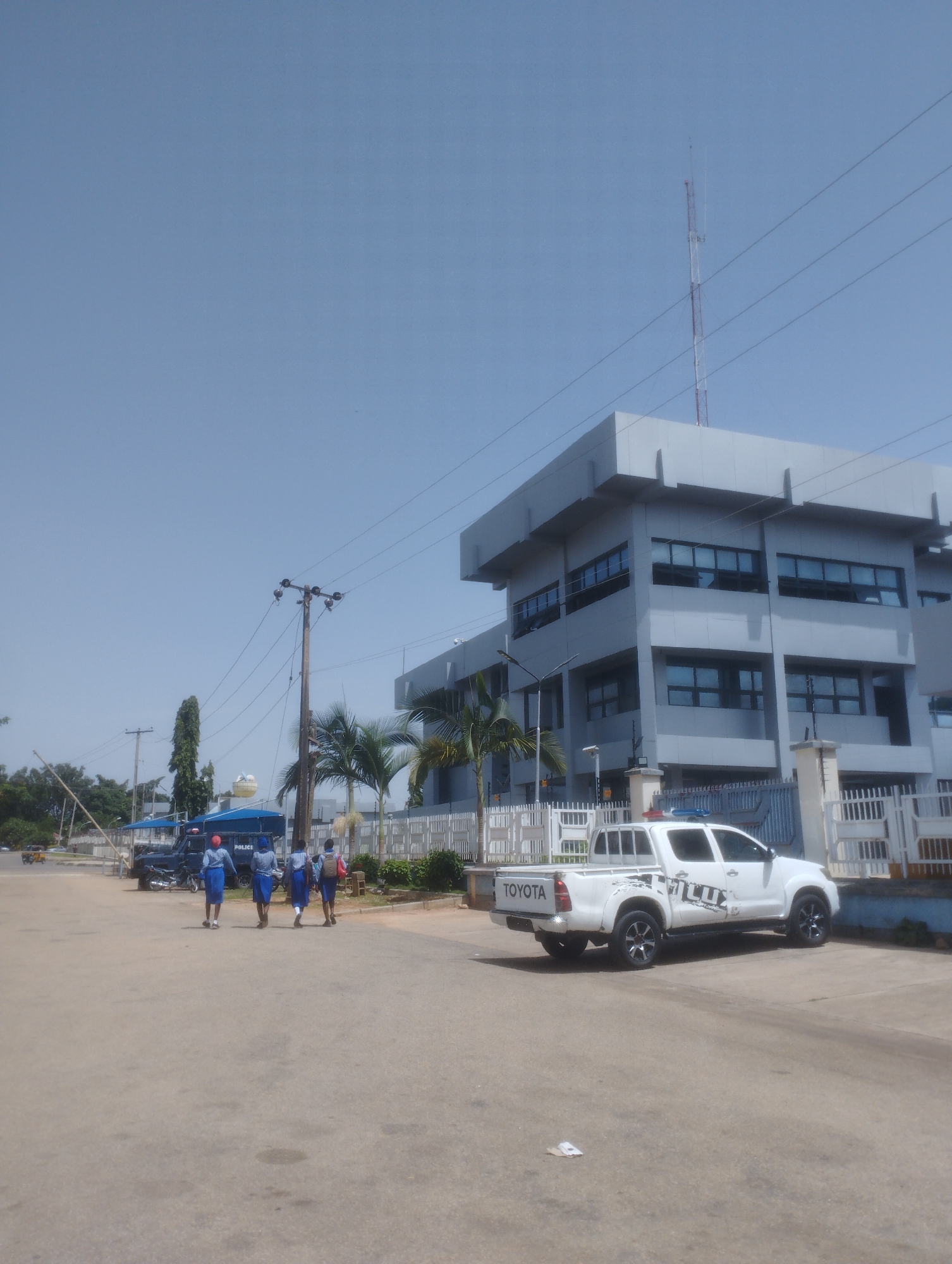 The image depicts an outdoor scene in the city of Jos, Nigeria. In the foreground, a paved road leads towards a multi-story building with a modern, grey facade and numerous windows. A white Toyota pickup truck, possibly a police vehicle given the lights on its roof and "POLICE" visible on a dark blue van in the background, is parked to the right of the road, facing away from the camera. Further left, a dark blue van with "POLICE" written on its side is parked near a gate. Three individuals, dressed in blue and white uniforms, are walking away from the camera on the road, towards the police van. The environment includes palm trees, power lines, and a tall communication tower in the distance against a clear blue sky. The overall scene suggests a governmental or institutional setting, possibly a police station or administrative complex.