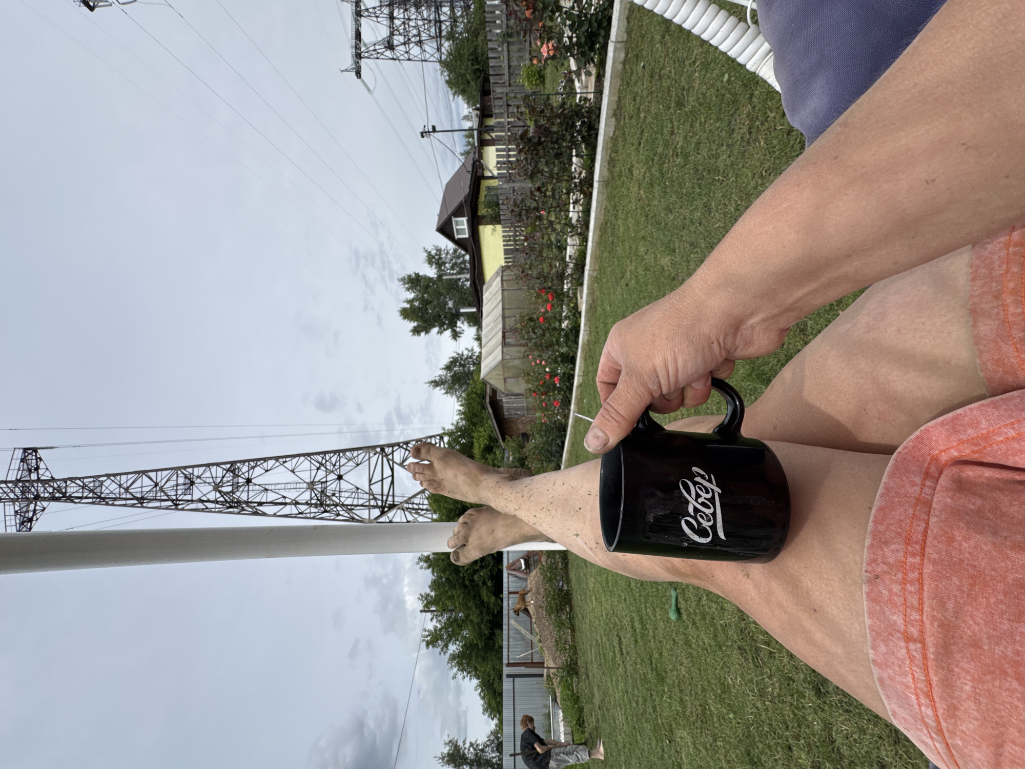 The image depicts a person's legs and arms resting in what appears to be a yard with a swing chair. The person is holding a black mug with the word "Север" written on it in white cursive. The background shows a grassy area with a fence, a greenhouse, a house, and tall electrical towers. In the distance, a person is seen working in the yard, and a dog is present. The sky is overcast. This scene is consistent with a residential backyard in the city of Chaikovsky, Russia, possibly during summer or a mild season, given the outdoor setting and the presence of greenery and flowers.