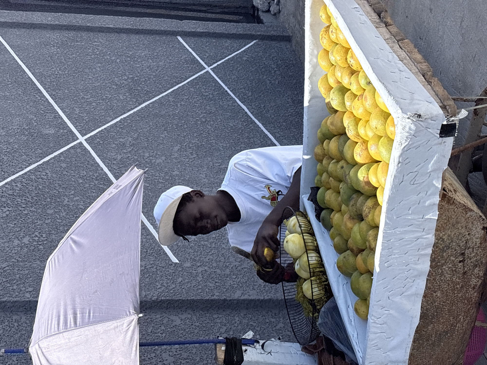 A man in Igboefon, Nigeria, stands by a fruit cart laden with yellow and green citrus fruits. He wears a white t-shirt and cap and appears to be cutting a fruit. Beside the cart stands a white umbrella. The background shows a gray concrete surface with white markings.
