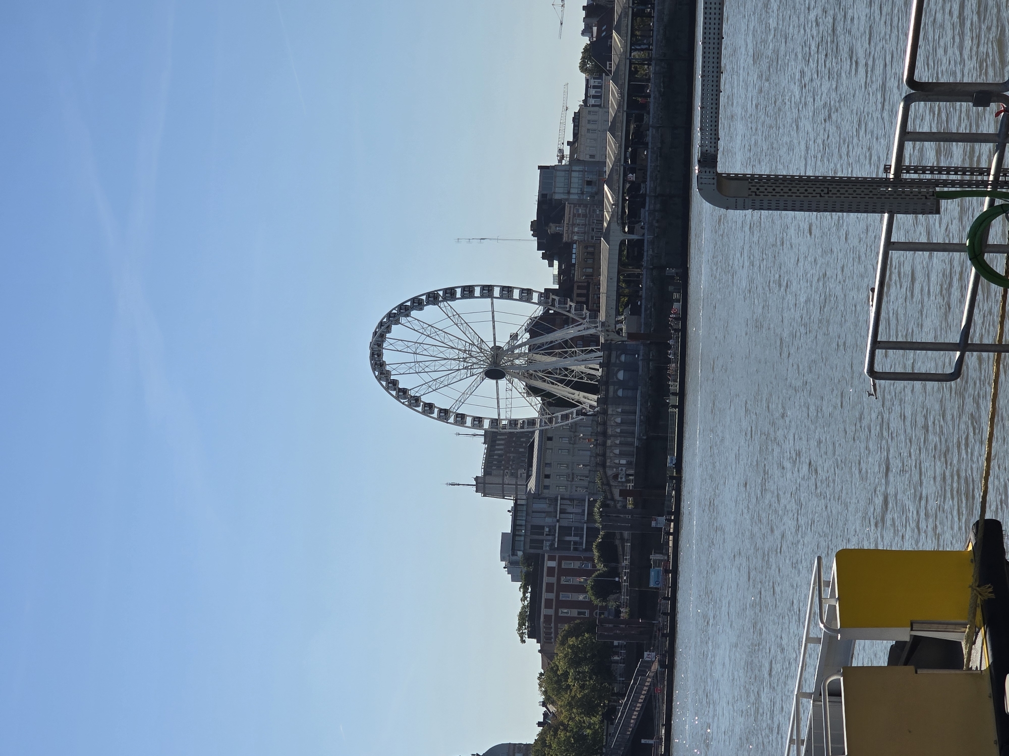 The image shows a sunny day in Antwerp, Belgium, with the large Ferris wheel of the Antwerp 360 visible in the background. The Ferris wheel is white and stands out against the clear blue sky. In the foreground, a body of water, likely the River Scheldt, reflects the sunlight. Part of a boat or ferry is visible in the bottom left and bottom right, with railings and yellow structures. The middle ground features the Antwerp skyline, with various buildings of different architectural styles, including some with red brick and others with modern glass facades. Trees are also visible, adding a touch of greenery to the urban landscape. A walkway or pier extends along the waterfront, with people visible on it. The overall scene depicts a vibrant city environment with a major attraction.