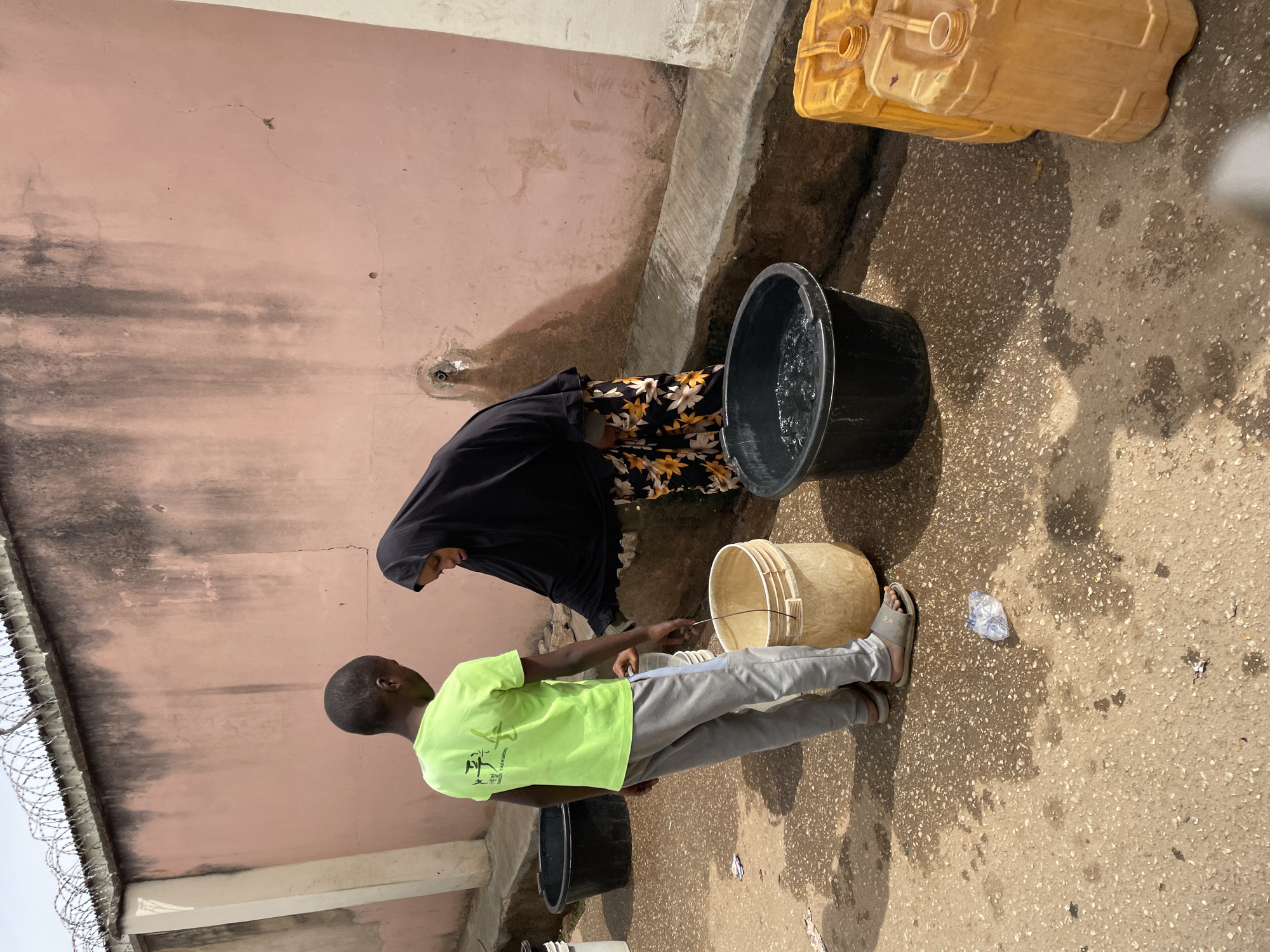 Two people, a woman and a boy, are standing near a pink exterior wall of a building. The woman is wearing a black hijab and a floral patterned dress, bending slightly towards a large black bucket filled with water. The boy, wearing a lime green t-shirt and grey pants, is holding a beige bucket. Two yellow jerry cans are positioned near the wall behind the black bucket. Additional black buckets are visible. The scene is in Jos, Nigeria.