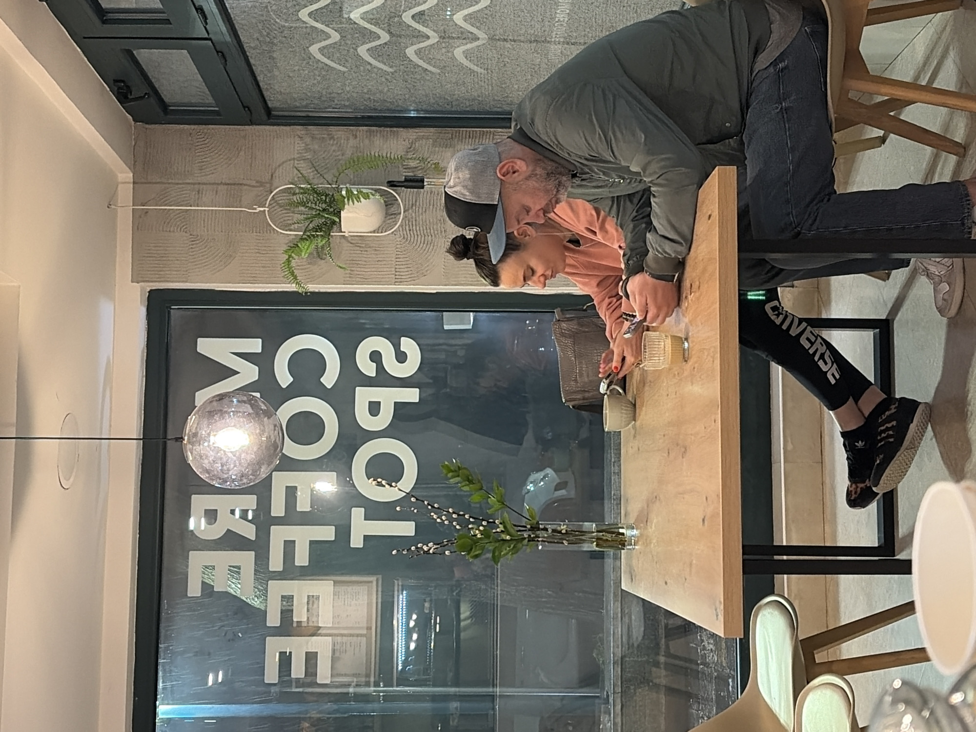 The image depicts an indoor scene at the More Coffee Spot cafe in Budva, Montenegro. Two adult individuals are seated at a light-wood rectangular table with black metal legs.
The male subject, on the right side of the table, wears a dark grey/olive green bomber jacket over a grey t-shirt, dark blue jeans, and a two-tone baseball cap (grey front, black back). He has a beard and is holding a smartphone with both hands, looking down at the screen. His feet, wearing light-colored sneakers, are visible beneath the table.
The female subject, on the left, wears a peach-colored long-sleeved top and dark leggings with "CONVERSE" lettering vertically on the left leg. Her hair is styled in a bun, and she appears to have wired earbuds. She holds a smartphone, looking down, and displays red nail polish on her fingernails. Her feet, wearing black patterned sneakers with white soles, are visible.
On the table, a tall, clear glass vase containing branches with green leaves and small white buds stands centrally. To its right, a light-colored ceramic cup is present. A textured glass with a light-colored beverage is positioned in front of the male subject. A brown handbag is partially visible near the female subject.
The background features a large window on the left, displaying the reversed white lettering "MORE COFFEE SPOT" and reflections of the interior. To the right of the window, a light-colored wall with a textured pattern of concentric circles is visible. A white hanging planter with a green fern is suspended against this textured wall. A spherical clear glass pendant light with an illuminated bulb hangs from the light-colored ceiling. The overall lighting suggests an evening or night setting. Two light-colored, wood-framed chairs are partially visible.