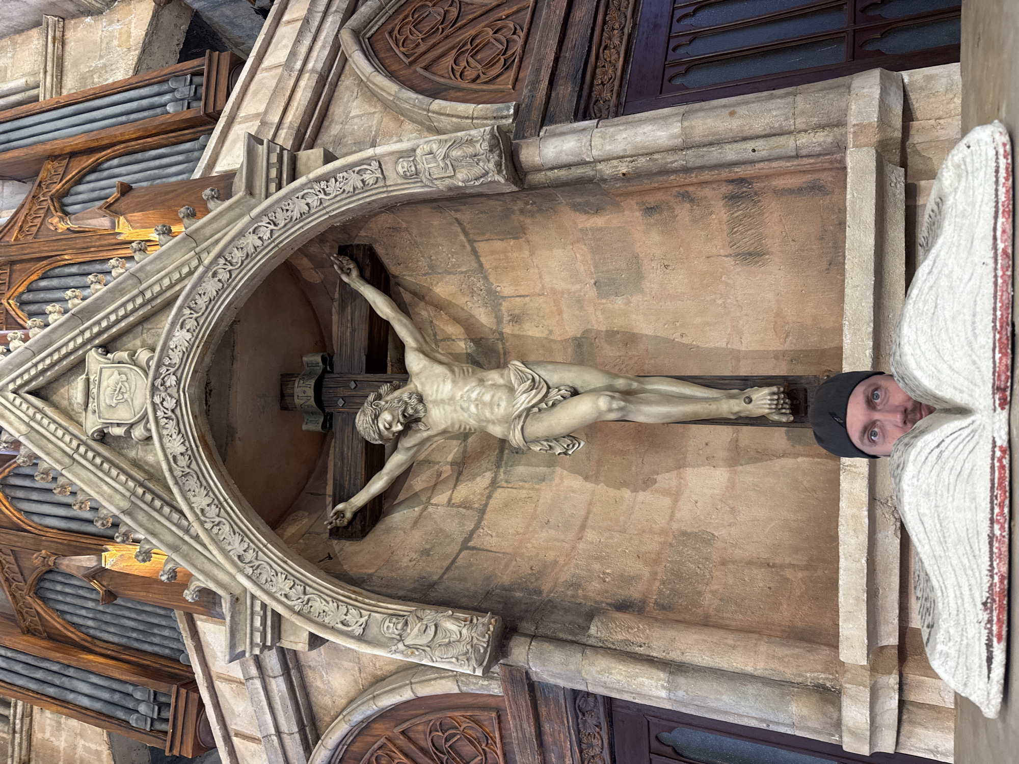 The media file captures the ornate interior of what appears to be a church or cathedral, located in Đà Nẵng, Vietnam, featuring prominent Gothic-inspired architectural elements. At the center, a large, light-colored sculpture of Jesus Christ hangs on a dark wooden crucifix, enshrined within an intricately carved stone archway. The archway is adorned with detailed foliage patterns and small decorative corbels or figures; one visible carving depicts a figure holding a book.
To the right, an adult male, wearing a dark cap, peers from behind a large, white, textured architectural element, looking directly towards the viewer with a neutral expression. His body is largely obscured.
The background reveals more of the building's rich detailing, including wooden structures with visible pipes, suggesting a large pipe organ on the upper left. Dark wooden panels with carved rosettes and other motifs are also visible, along with dark window frames in the upper sections. The stonework exhibits varied textures and warm, earthy tones, from light beige to deeper browns, indicating age and craftsmanship. Near the head of the crucified figure, a small scroll or plaque bears some indistinct, illegible text. The lighting is even, suggesting an indoor setting without direct sunlight.