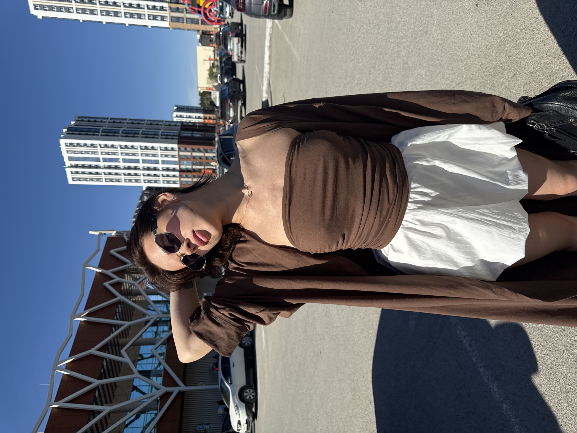 A woman is posing in a parking lot set against a backdrop of buildings and a clear blue sky. She is wearing sunglasses, a brown top, a white skirt, and a long brown cardigan. Her head is tilted, and her tongue is slightly visible. In the background are several cars and modern architectural structures. Tall buildings, with repetitive window patterns, are also visible. The photograph appears to have been taken at a tilt. The location is Perm, Russia.
