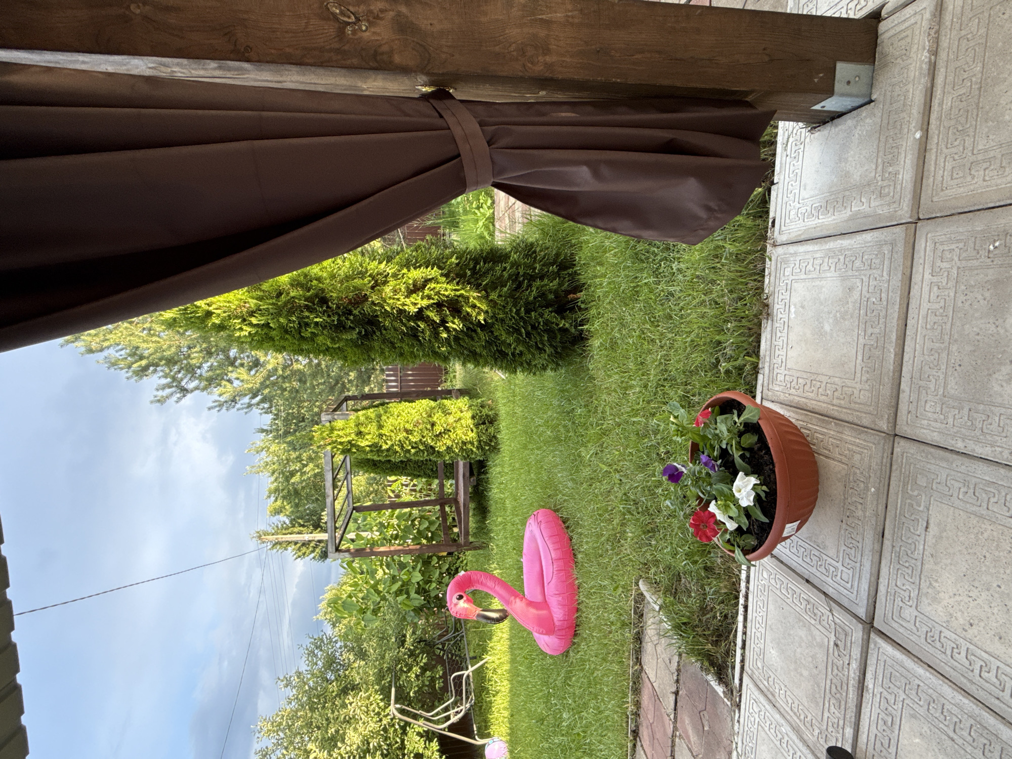 A bright pink inflatable flamingo float rests on a vibrant green lawn in a backyard. The flamingo is positioned near a swing set, with a wooden gazebo structure visible in the background. Two tall, slender evergreen trees frame the scene. In the foreground, a brown planter filled with colorful flowers sits on a stone patio. The sky above is partly cloudy, with a mixture of blue and grey. The overall setting appears to be a serene outdoor space in a suburban or rural environment, possibly within the city of Тмз Снт, Russia.