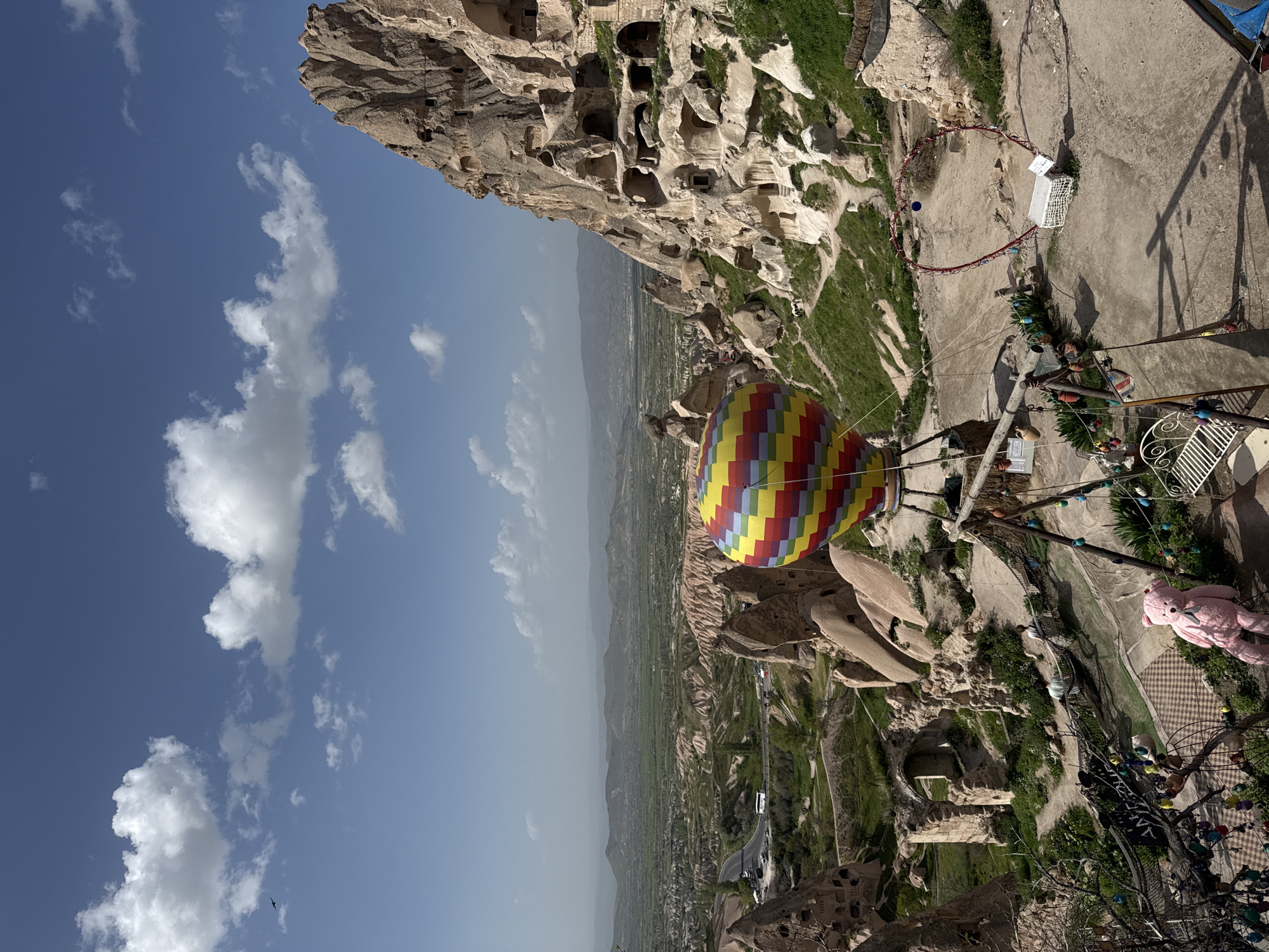 The image captures a scene in Uçhisar Beldesi, Turkey, featuring a multicolored hot air balloon suspended against a partly cloudy sky and the distinctive landscape of Cappadocia. The balloon, composed of red, yellow, blue, and green panels, is positioned near a cliff face marked with numerous cave dwellings carved into the rock. The geological formations of the region are visible, with eroded stone structures and green vegetation dotting the terrain. In the foreground, there are structures that seem to be part of an observation area or tourist spot, including a teddy bear, metal fences, and artificial decoration. The horizon reveals a vista of green fields stretching into the distance under a slightly hazy atmosphere.