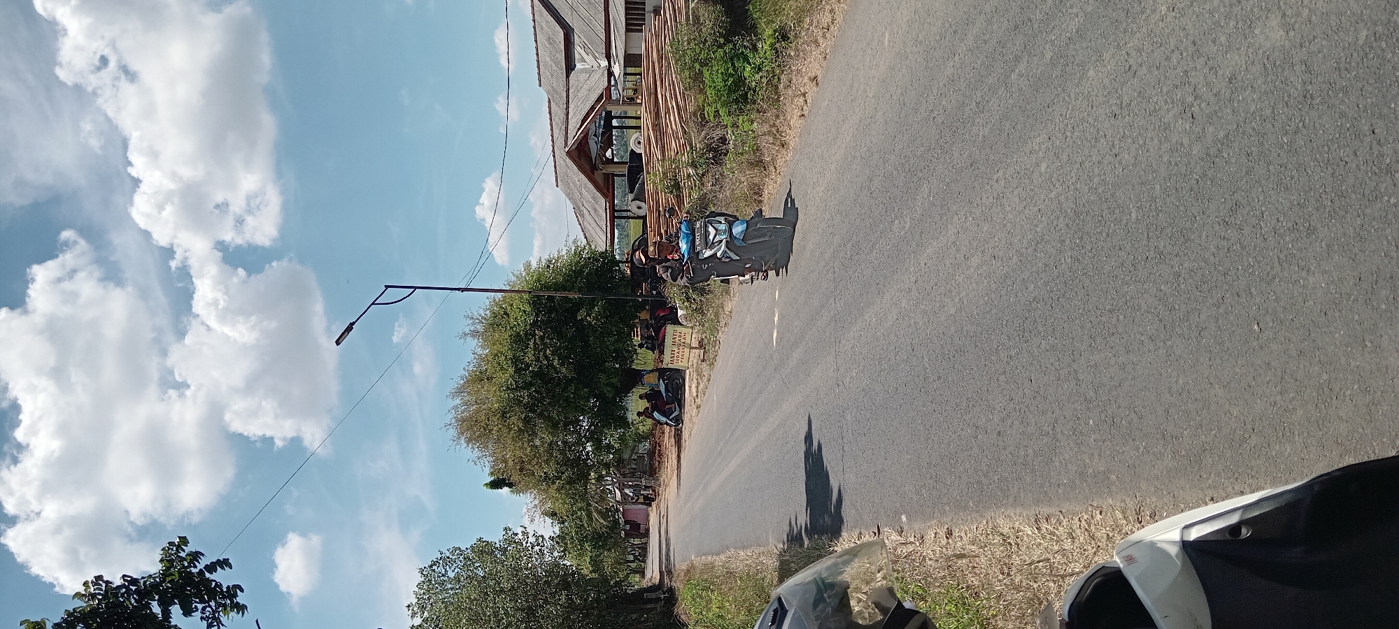 The image depicts a rural road scene in Paledah, Indonesia. A paved road stretches into the distance, bordered by dry grass and vegetation on the left and right. On the right side of the road, a building with a thatched roof is visible, with a large stack of wood or bamboo poles in front of it. Several motorcycles are parked near the building. On the left side of the road, a line of trees and small structures are present. In the foreground, the front of a white and black scooter is visible. Further down the road, a blue motorcycle is being ridden by a person wearing a hat. A street lamp stands on a pole on the right side of the road. The sky is blue with scattered white clouds. A yellow sign with Indonesian text "HATI HATI SEDANG ADA PEKERJAAN" meaning "BE CAREFUL, THERE IS WORK BEING DONE" is placed on the side of the road.