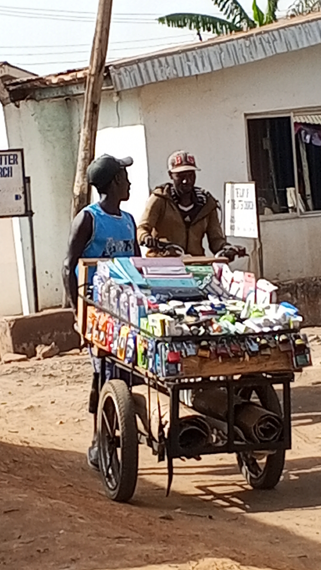 Two men stand next to a cart laden with merchandise in Sabon Tasha, Nigeria. The man on the left wears a blue tank top and a dark cap, while the man on the right wears a brown jacket and baseball cap. The cart is filled with various items, including what appears to be stationery, electronics, and other small goods. The cart also has wheels and a metal frame. Behind them are buildings, a wooden utility pole, and a sign. The ground is sandy and sunlit.