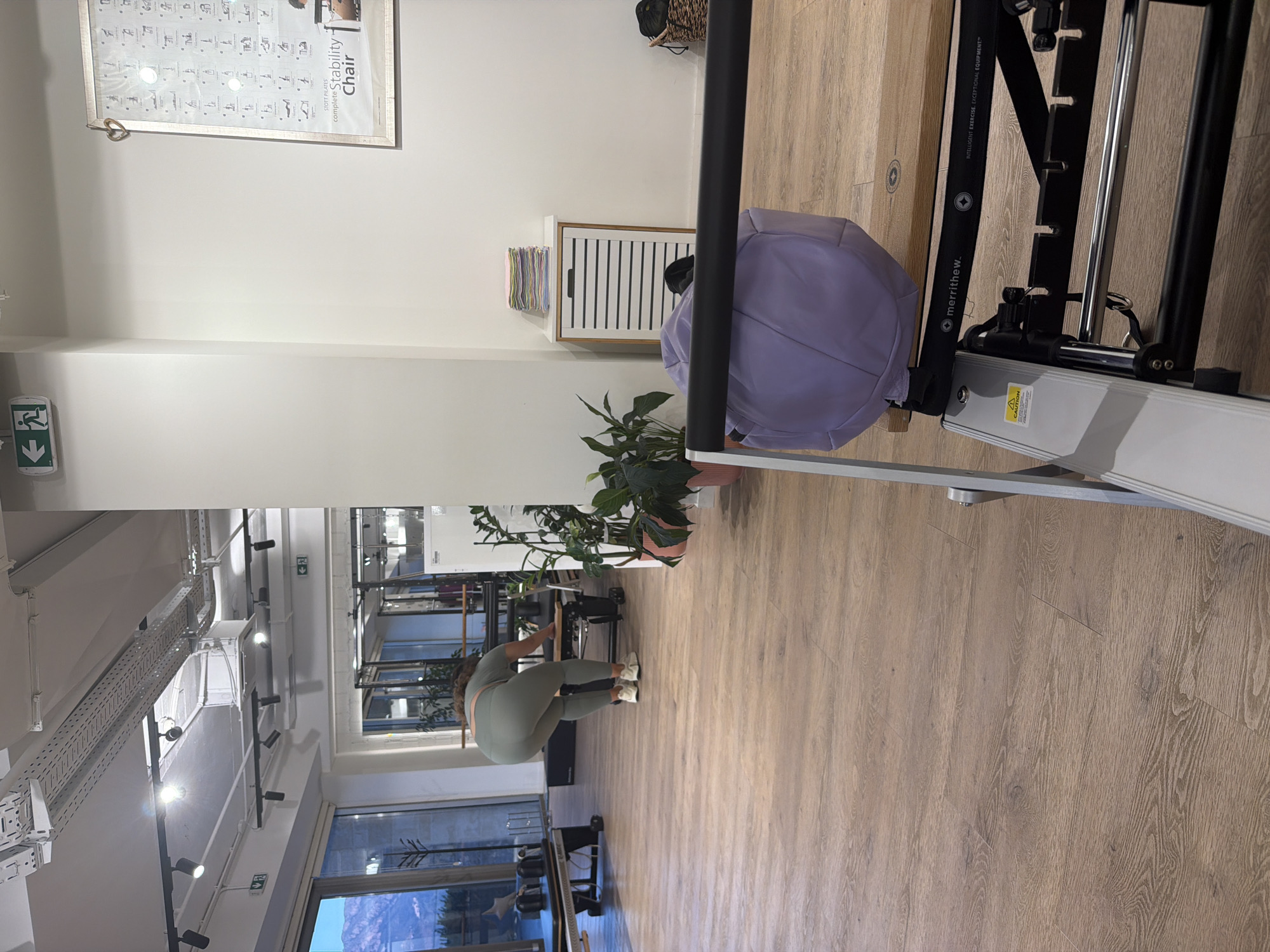 In a brightly lit indoor space, likely a fitness studio in Boreti, Montenegro, a woman in olive green athletic wear is captured from behind. She is bent over, her hands resting on a piece of equipment that appears to be a pilates reformer. The studio features light wood flooring and industrial-style ceiling fixtures. To the right, a white cabinet with horizontal slats is visible, topped with a stack of colorful folded towels. A large purple cushion rests on the reformer, and a black basket sits on a shelf in the background. A poster, potentially detailing pilates exercises, hangs on the far wall. The scene is devoid of any other individuals.