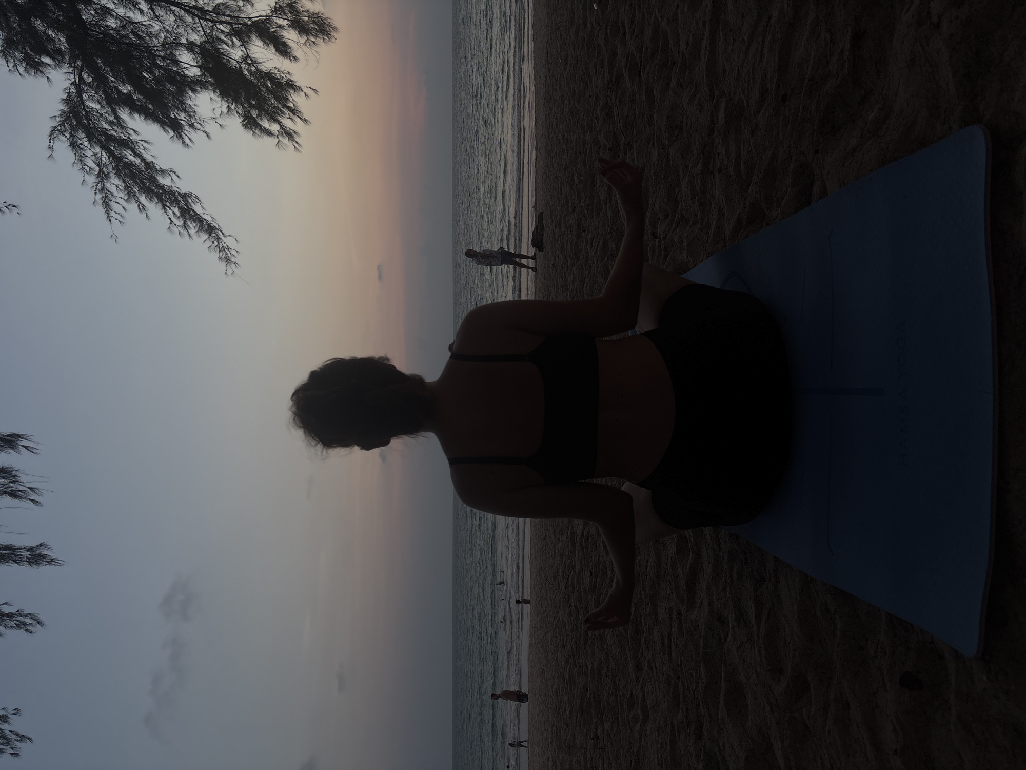 A person is doing yoga on a blue mat on a beach in Choeng Thale, Thailand. The person is facing the ocean, which is calm with a few people in the distance. The sky is a gradient of colors from blue to orange. There are tree branches in the upper left corner of the frame.