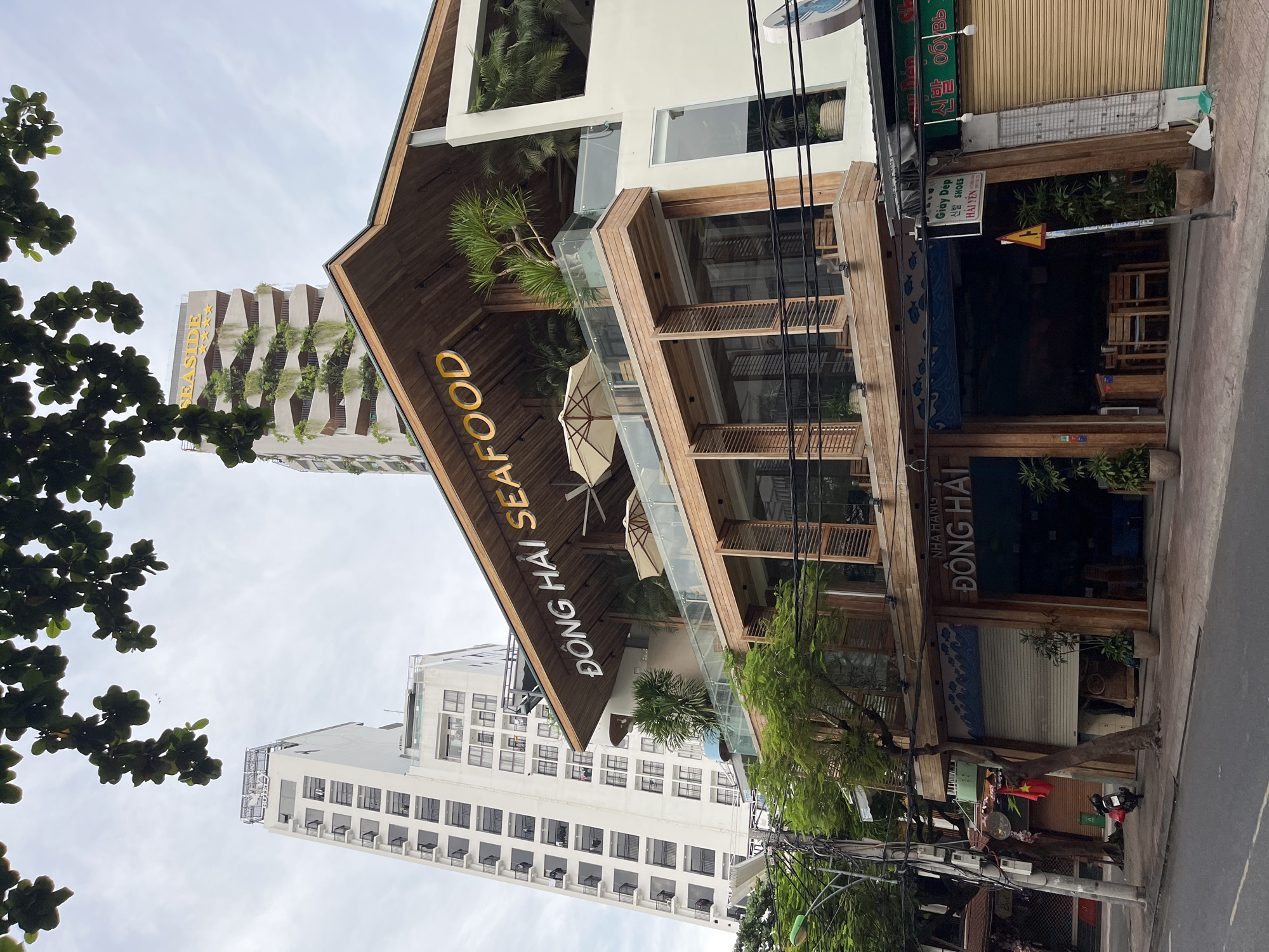 The image shows a multi-story building in Nha Trang, Vietnam, with a prominent seafood restaurant named "ĐÔNG HẢI SEAFOOD" on its upper levels. The restaurant features a modern design with wooden accents, glass railings, and outdoor seating areas with umbrellas. In the background, two tall white buildings, one appearing to be a hotel with the name "SEASIDE" visible on top, rise against a cloudy sky. Green foliage from trees and plants adorns the restaurant's balconies and the surrounding environment. The ground level of the building displays a closed shop with a rolled-down shutter, a Vietnamese flag, and a parked motorbike. A street with a sidewalk and overhead power lines is in the foreground, indicating an urban setting. There are no people visible in the immediate scene.