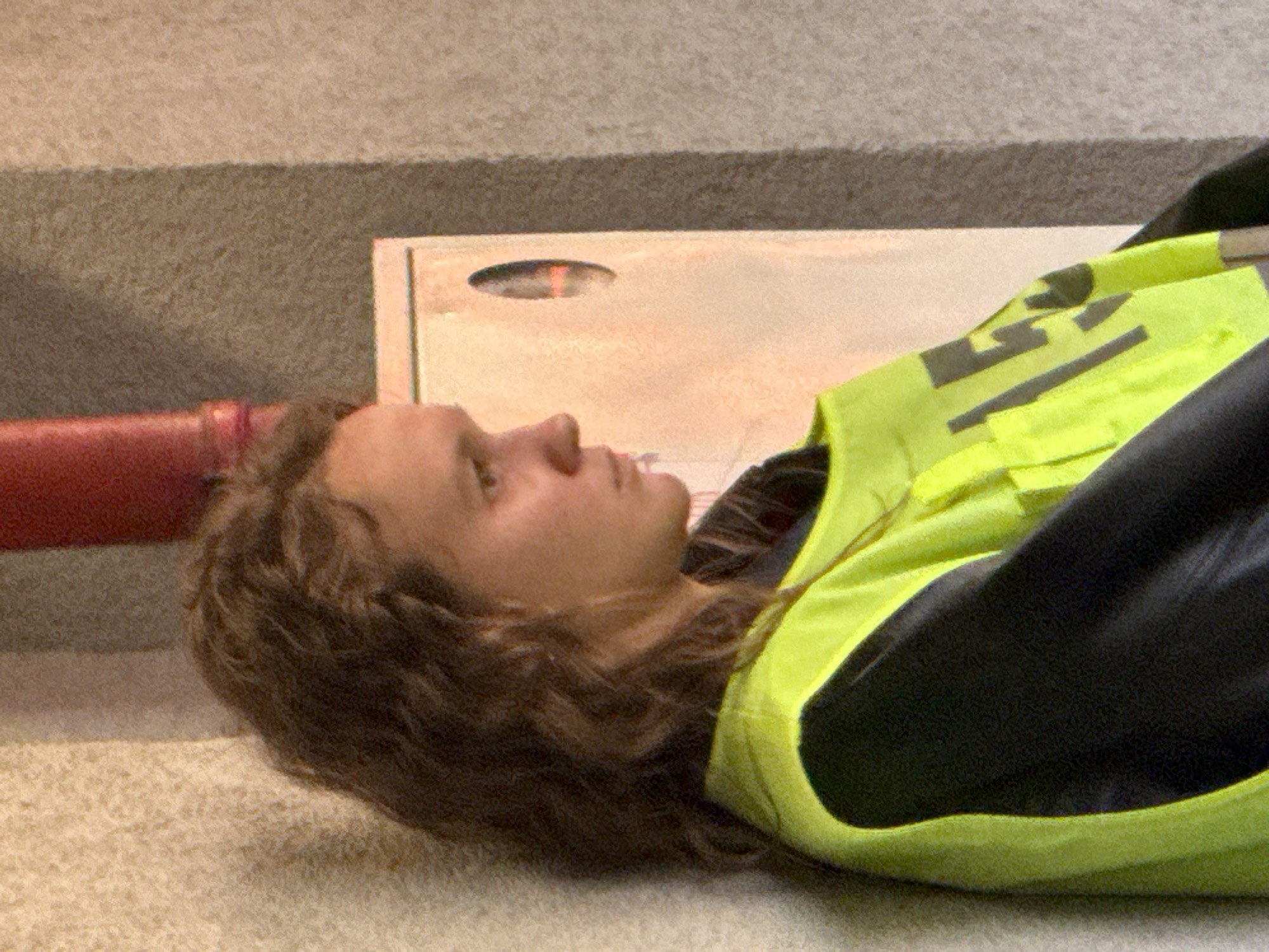 The image shows a person lying on the ground near a wall and what appears to be a fire hose cabinet. The person has light brown hair and is wearing a black jacket and a fluorescent yellow vest with dark markings. The background includes a reddish-brown fire hose and a textured wall. Without additional context or information, it is not possible to specifically identify the location within Yekaterinburg, Russia. The scene appears to depict a staged or reenacted scenario, given the individual's prone position and the proximity to a fire safety device.