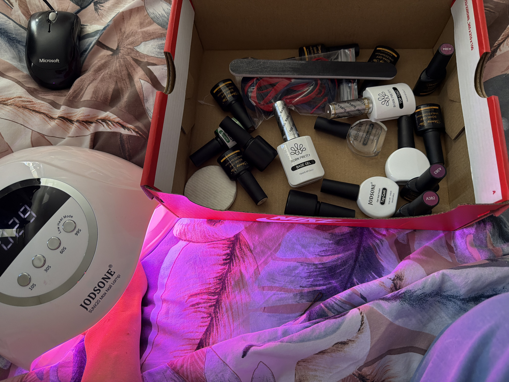 The image captures a casual, indoor setting in Bearpark, United Kingdom, featuring a collection of nail care essentials and a computer mouse on a patterned fabric.
A white JODSONE SUNX20 Max Nail Lamp occupies the lower left, emitting a distinct pink-purple light. Its digital display shows "29" (likely seconds remaining), and it has buttons for "10S", "30S", "60S", and "99S" (Low Heat Mode). An open cardboard box, marked with "52-22N5W8 265x210x100" on its side, is filled with various gel nail polish bottles and tools. Brands visible include "BORN PRETTY" (with "BASE GEL" and "TOP COAT"), "Shining She" ("SOAK-OFF UV&LED Gel Polish"), "JODSONE" ("Gel Nail Polish Top Coat" and "Nail Dehydrator"), and "JIAZHIGAI". Some bottles are labeled with color codes like "A382", "A383", and "A384". A black nail file, red string or band, and white cotton pads are also scattered within the box.
In the upper left, a black wired Microsoft computer mouse rests on the fabric. The entire scene is laid out on a soft, patterned fabric, possibly bedding or a blanket, adorned with abstract floral or leaf designs in shades of pink, purple, brown, and cream. The presence of the nail lamp and numerous polishes suggests a home manicure or nail art session is either underway or has recently concluded.