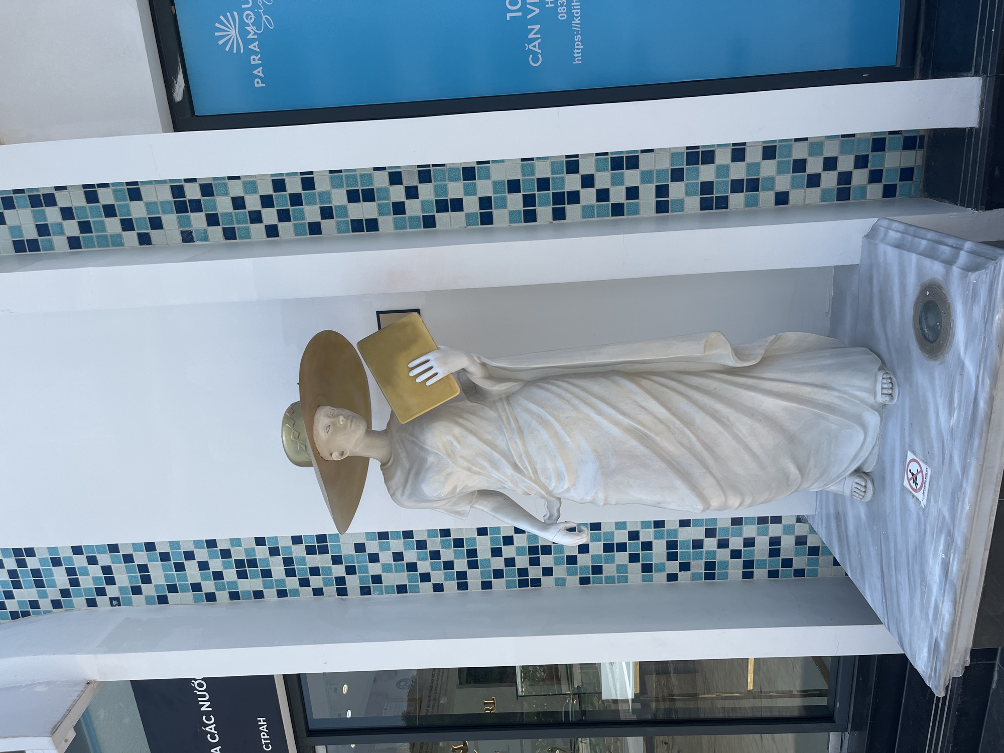 A white marble statue of a woman wearing a large, wide-brimmed hat stands on a marble pedestal in front of a building with a tiled facade. The statue is dressed in a flowing, draped garment and holds a gold-colored book in her right hand. The building's facade is decorated with a mosaic of blue and white tiles. To the left of the statue, a glass storefront displays merchandise. To the right, a blue sign with white text reads "PARAMOUNT." The statue is positioned outdoors, likely on a sidewalk or in a public square. A small, circular sign with a prohibition symbol is visible on the pedestal near the statue's feet. The scene appears to be a street in Nha Trang, Vietnam.