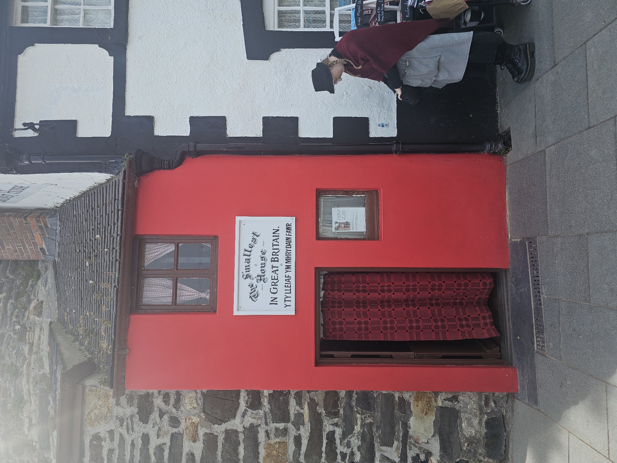 A medium shot reveals "The Smallest House in Great Britain" nestled in the city of Llandudno Junction, United Kingdom. The tiny structure is painted a vibrant red. A woman with a black hat is standing outside of the building. Next to the house on the left is a stone wall, and on top is a shingled roof. Directly in front of the house is an entry space with a brown frame and a red checked fabric inside. A window is to the left of the entryway, and another, smaller window is to the right. A sign to the left of the door reads, "The Smallest House In Great Britain. Y TY LLEIAF YM MHRY DAIN FAWR." The building next to the house is white and black. The street is to the right of the house.