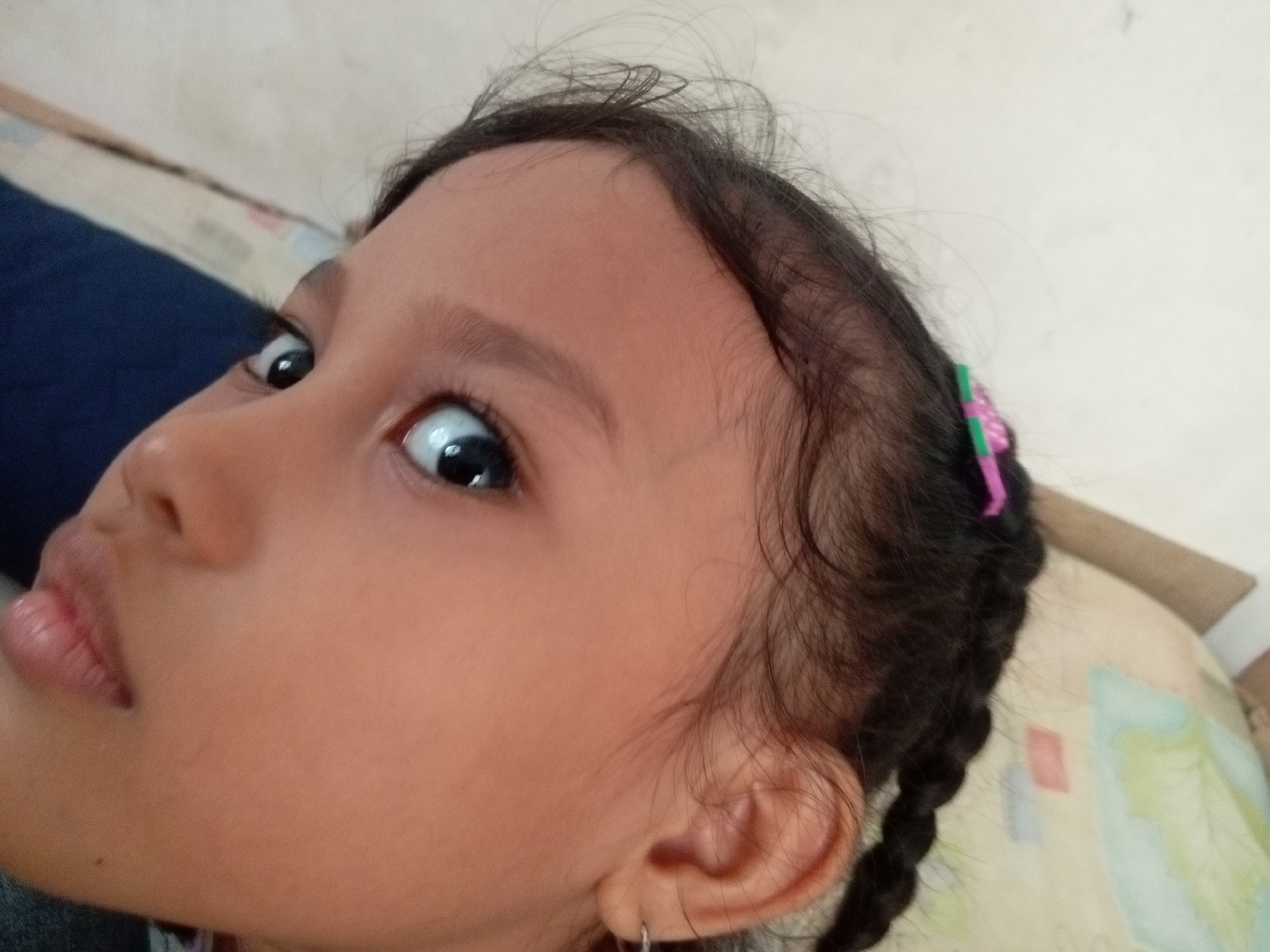 A close-up, high-angle shot shows a young girl's face, with her left eye and mouth prominently featured. The girl has dark hair, which is braided and adorned with a pink and green hair accessory. She is wearing a silver hoop earring in her left ear. Her skin tone is light brown, and her eyes are wide open, looking upwards and to the left. The background is blurred, but appears to be a light-colored surface, possibly a bed or couch with a patterned textile. The image is a portrait of the girl's head and upper face. The metadata indicates the image was taken in Batam, Indonesia.