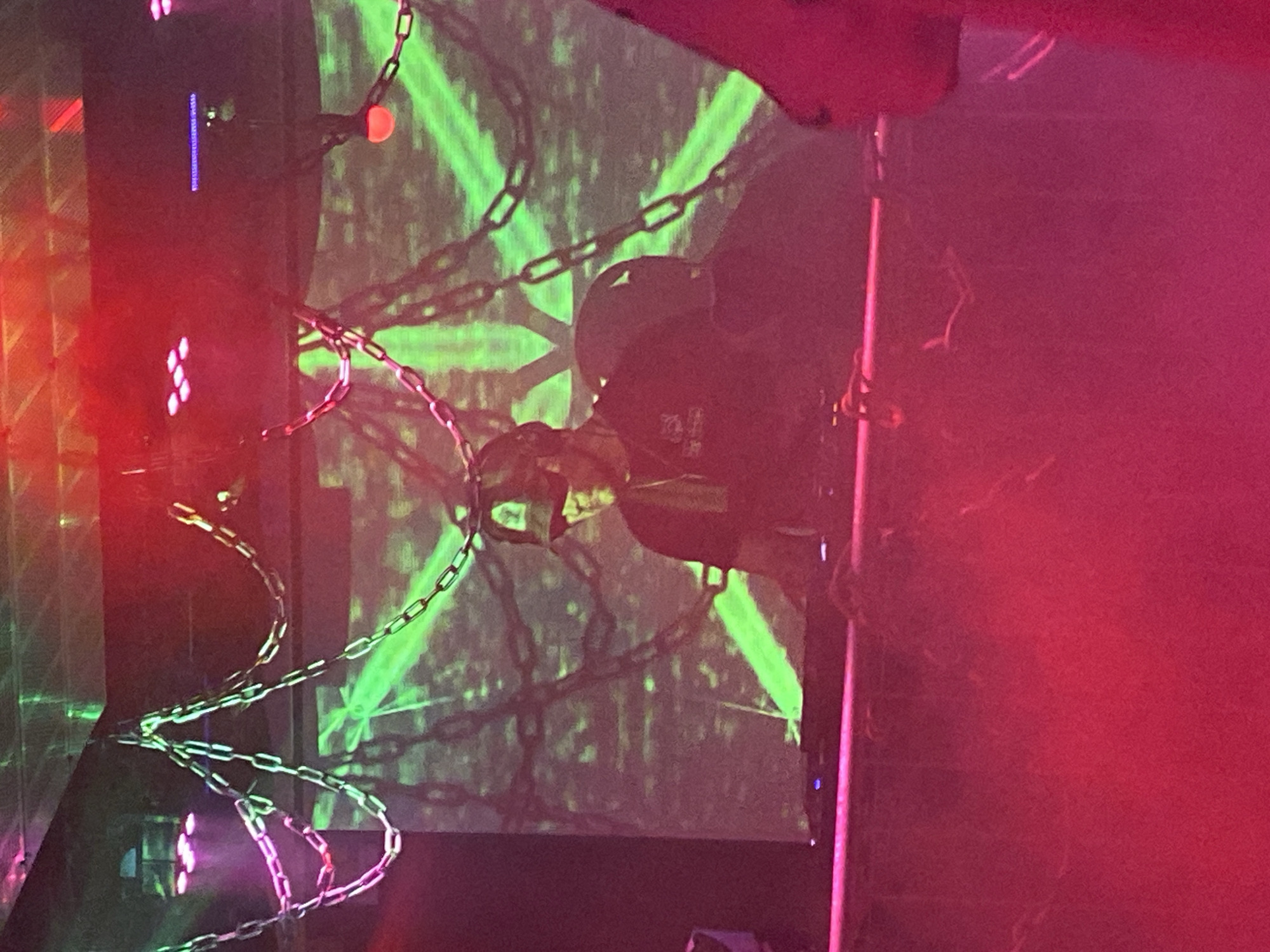 A DJ is performing at a nightclub in Joensuu, Finland. The DJ is wearing a cap and a dark shirt, and is standing behind a DJ booth. There is a large projection screen in the background with a green star pattern. The ceiling is decorated with chains and lights. The atmosphere is dark and smoky, with red and green lighting.