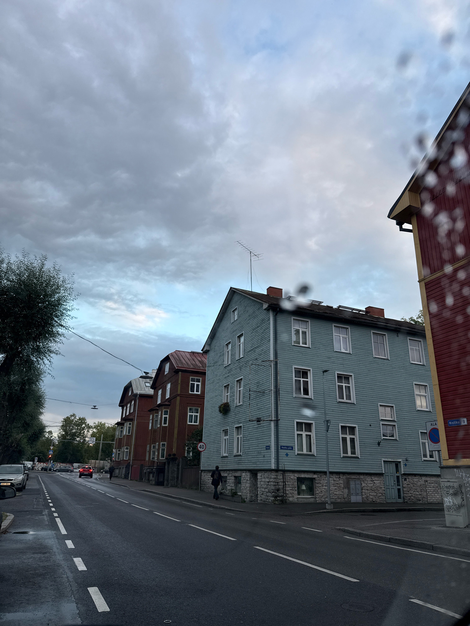 The image shows a street scene in Tallinn, Estonia, with residential buildings lining the road. The sky is overcast, with heavy clouds suggesting an impending rain or recent rainfall, as evidenced by the water droplets on the camera lens in the foreground.
On the right side of the image, a red-and-yellow building with a dark roof is partially visible, featuring an overhang and a downspout. To its left is a light blue, multi-story building constructed with horizontal wooden siding and a stone foundation. This building has numerous white-framed windows and a dark-colored door on the ground floor. A street sign indicates "Ristiku 9" and "ALA," suggesting this is Ristiku Street. A circular speed limit sign with "40" is also visible.
Further down the street, to the left of the blue building, are several red brick buildings with pitched roofs and white-trimmed windows. These appear to be residential homes.
The street itself is asphalt, with white dashed lane markings. Several cars are present on the road, including a silver sedan parked on the left side of the street and a red car further down. Two figures are walking on the sidewalk in front of the buildings, one near the blue building and another further down the street. A large tree with dark foliage stands on the far left of the image.
The overall atmosphere is muted, characteristic of an overcast day, and the presence of water droplets on the lens adds to the impression of wet weather.