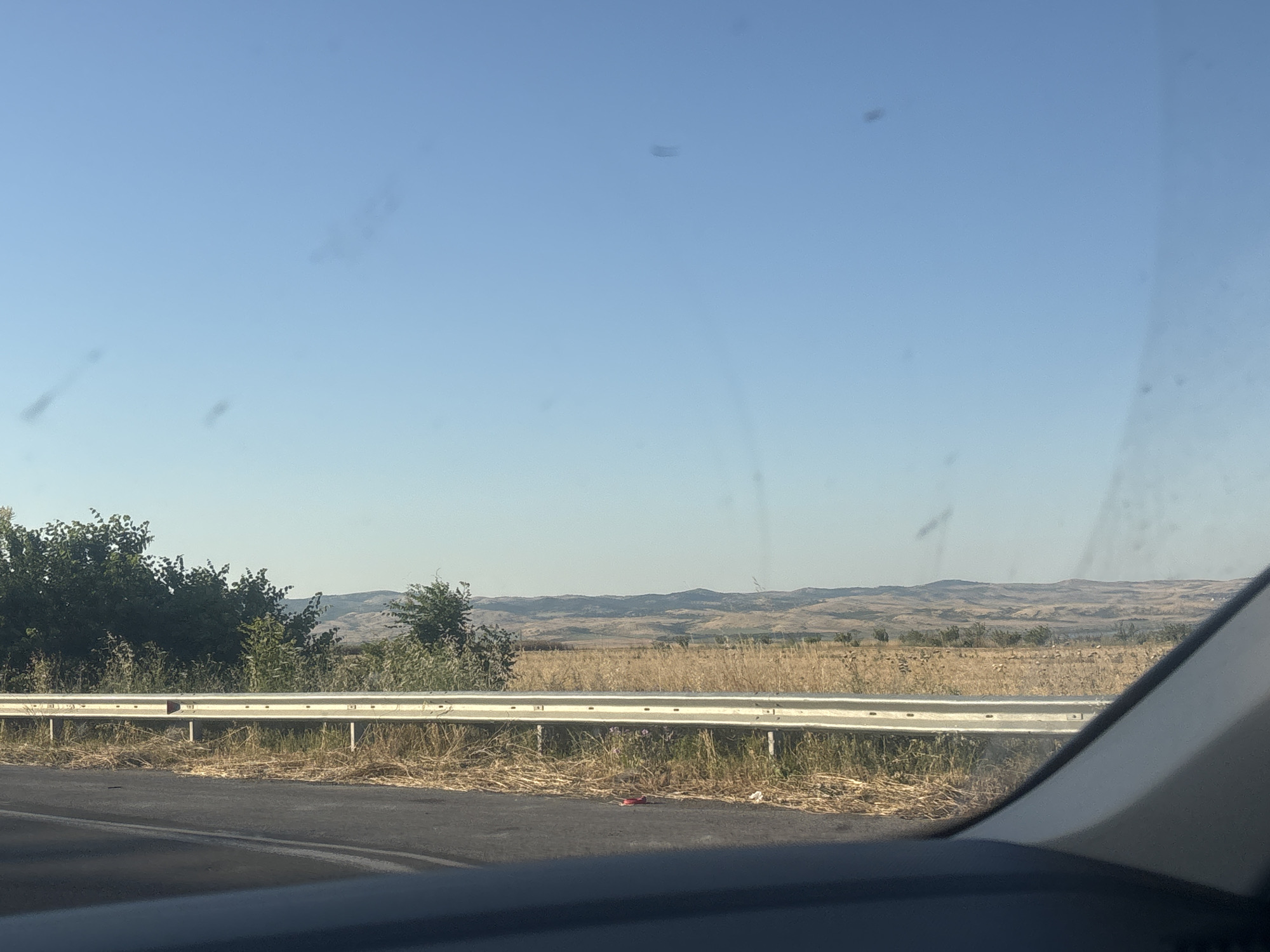 The media file depicts a view from inside a vehicle traveling along a road, likely in Burgas, Bulgaria. The scene shows a clear, bright blue sky with a few specks of dust or debris on the windshield. A silver guardrail runs along the side of the road, separating it from a dry, grassy field. In the background, rolling hills are visible under the hazy sunlight, suggesting it is daytime and likely a warm, dry season. Some green trees and bushes are scattered along the edge of the field and on the hills. The overall impression is one of a rural landscape seen from a moving vehicle. There is no text visible in the image.