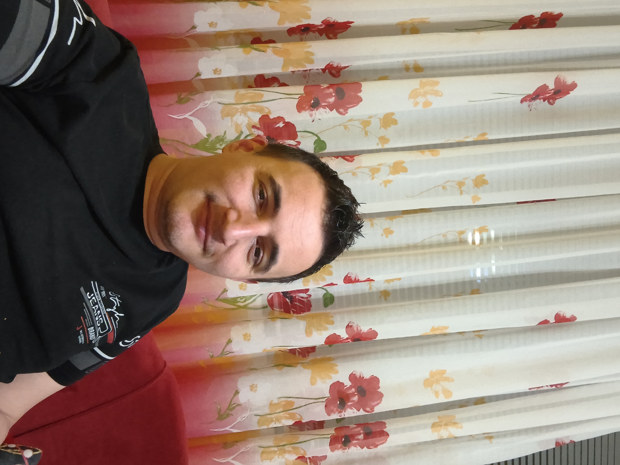 A male individual, appearing to be an adult, is depicted seated indoors against a floral curtain and a red upholstered couch. The man has dark, spiky hair, light skin, and a faint mustache and chin stubble. He is smiling subtly and looking directly at the camera. He wears a black short-sleeved t-shirt with a white and grey graphic design on the chest, which includes the text "JEANS BRAND" and "Style".
The background features white sheer curtains adorned with a repeating floral pattern. The pattern consists of prominent red poppy-like flowers with green stems, interspersed with yellow and some white floral designs. The lower portion of the curtains transitions into a reddish-pink hue. Faint horizontal blinds are visible through the sheer fabric of the curtains in some areas. A portion of a red couch or armchair is visible on the right side and bottom right corner of the image, serving as the seating for the individual. The setting appears to be an interior domestic space. While the image itself does not provide explicit visual cues for a specific geographic location, the context implies the city of Shumen, Bulgaria.
