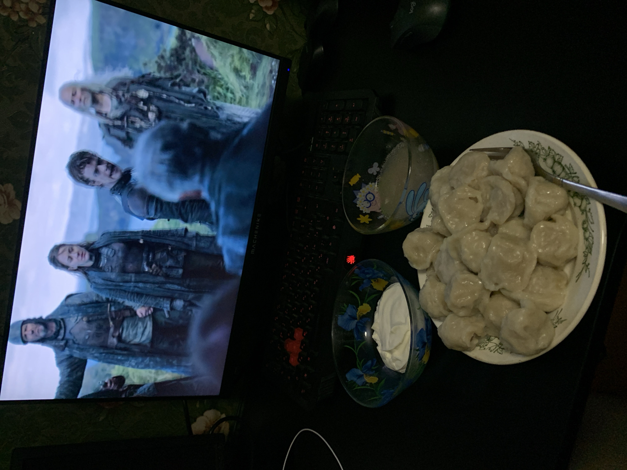 A close-up, eye-level shot captures a dimly lit scene featuring a plate of dumplings, two small bowls, a computer monitor displaying a medieval-themed scene, and a keyboard. The plate, positioned centrally in the foreground, is filled with numerous light-colored dumplings arranged in a circular pattern. To the left of the plate, a bowl contains a dollop of white cream, possibly sour cream or yogurt. To the right, another bowl holds a light-colored liquid. The computer monitor, set further back, shows a screenshot from a show or movie with several individuals in historical attire. A keyboard with red-accented keys is visible beneath the monitor. The overall setting appears to be a home or office environment, given the presence of a computer and desk. The image does not provide any specific details related to the city of Desyatikhatka, Russia.