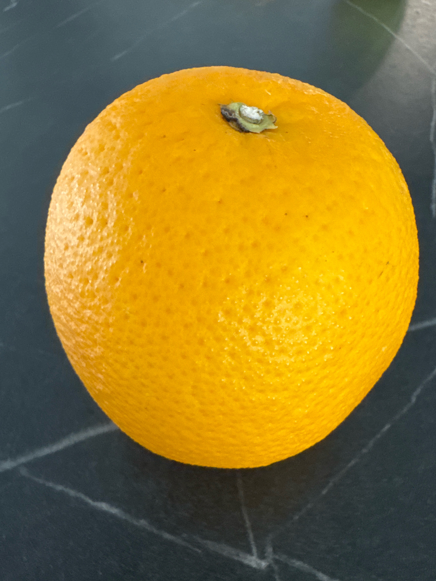 A single, whole orange sits on a dark, speckled countertop. The orange is bright orange with a textured rind and a small, green stem at the top. The countertop appears to be made of dark granite or a similar material with lighter, thin veins running through it. The lighting suggests an indoor environment, possibly a kitchen. The location is specified as Бараўлянскі сельскі Савет, Belarus.