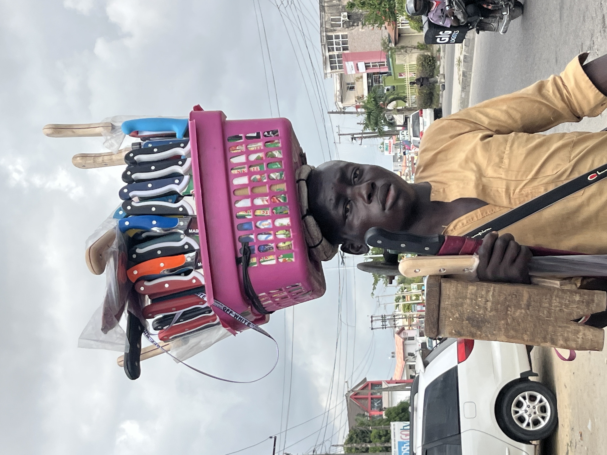 A portrait-oriented image, rotated 90 degrees clockwise, captures a street hawker in Ado, Nigeria, presenting his wares on an overcast day. The central figure is a young Black man, dressed in a light brown long-sleeved shirt, who looks directly at the camera with a neutral expression.
He skillfully balances a bright pink plastic laundry basket on his head, which appears to contain various packaged items, although their specific nature is largely indistinguishable. A white strap with the text "OFF WHITE" is visible, hanging from the basket. Attached around the basket and suspended downwards are numerous kitchen knives of varying sizes, seemingly packaged for sale. These knives feature diverse handle colors, including natural wood, blue, black, orange, and red. In his visible hand (his right, viewer's left), he holds a wooden block to which several larger knives or machetes are secured, also part of his merchandise.
The scene unfolds on an urban street. A paved road occupies the background, with a white car (possibly an SUV or minivan) parked on the side. Various buildings, painted in shades of red, green, and grey, line the street. Overcast clouds fill the sky, and a dense network of utility poles and power lines crisscrosses the frame. In the mid-ground, a person on a motorcycle with "GIG LOGISTICS" prominently displayed on its storage box is visible. Another distant sign partially reads "IMOT PHARMACY," suggesting commercial activity. The image portrays a common sight of street vending in Nigerian urban landscapes.