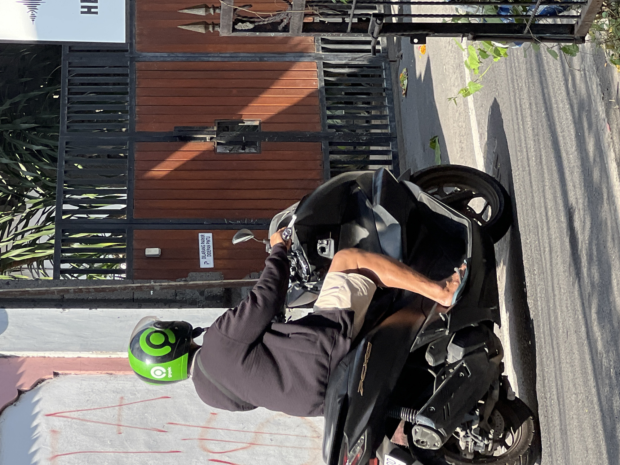A person wearing a green and black Gojek helmet is riding a black motorcycle. The rider is wearing a dark long-sleeved shirt and light-colored shorts, with their legs exposed and feet wearing flip-flops. The motorcycle is on an asphalt road, with a white line visible on the road. In the background, there is a textured wall with red markings, a dark metal gate with wooden panels, and foliage. A sign on the gate reads "DILARANG PARKIR DIDEPAN PINTU," which translates to "NO PARKING IN FRONT OF THE DOOR." There are also offerings on the ground near the gate. The scene appears to be in Kuta, Indonesia, indicated by the Gojek helmet, a ride-hailing service popular in the country, and the Indonesian text on the sign.