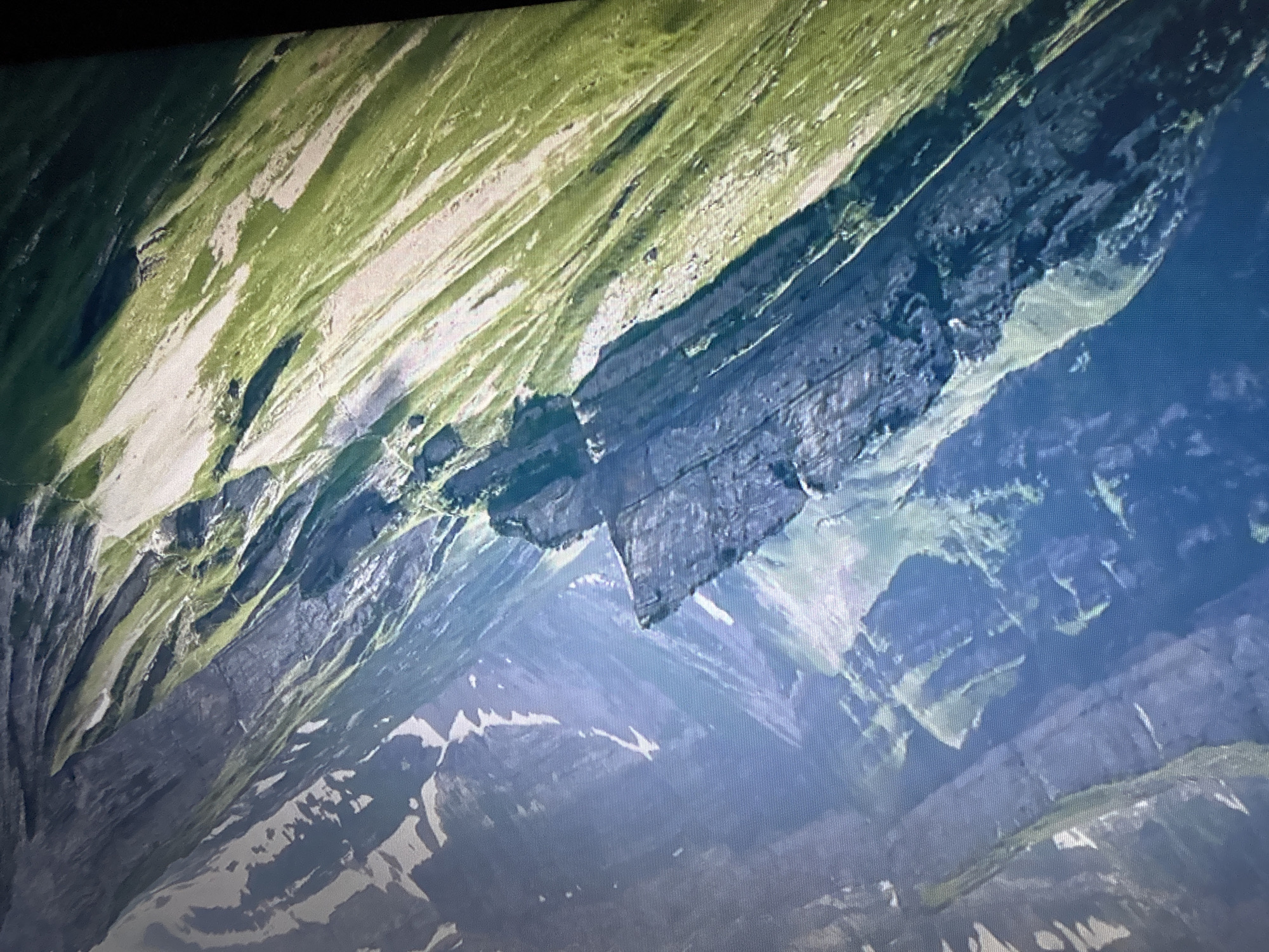 The image is a high-angle, close-up shot of a mountainous landscape. The mountains are steep and rugged, with sheer rock faces interspersed with slopes covered in lush green grass. Patches of snow are visible on some of the higher peaks and crevices, indicating a high altitude environment. The lighting suggests it is daytime, with sunlight casting shadows that highlight the textures and contours of the terrain. The overall impression is one of wild, untamed natural beauty. The location is not identifiable as being within any city.