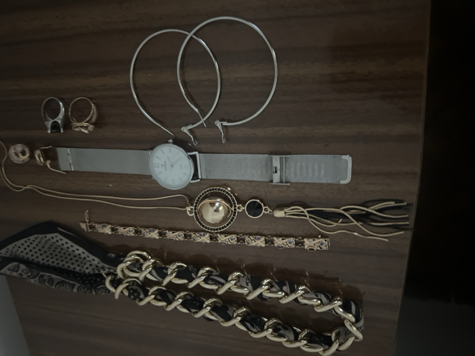 The scene is located in the city of Smederevska Palanka Municipality, Serbia.
An assortment of fashion accessories is displayed on a dark brown, reflective, polished wooden surface. From left to right:
On the far left, a folded fabric item, possibly a scarf or headband, features a black and beige/white geometric or paisley pattern with a distinct black border. Adjacent to it, a large, gold-toned chain accessory, likely a necklace or bracelet, has black fabric interwoven through its links. To its right, a gold-toned link bracelet is visible, accented with small, dark blue or purple-colored stones set within rectangular links.
Further to the right, a long gold-toned necklace is laid out, featuring a complex pendant composed of a spherical gold-toned element encased by a ring of black stones, followed by a smaller black circular disc, and terminating in a tassel made of gold and black strands. Immediately to the right of the necklace, a wristwatch with a silver-toned mesh band and an open clasp is visible. The watch face is white with black numerals or markers and displays the brand "CEL MARE".
Above the watch, two rings are present: one gold-toned ring with a patterned band and another silver-toned ring featuring a black rectangular inlay. To the right of these, a pair of large, silver-toned hoop earrings are laid flat. Below these, two individual small silver-toned earring clasps or hooks are also present.
No individuals are visible in the scene. The objects are stationary, with no actions or interactions depicted.