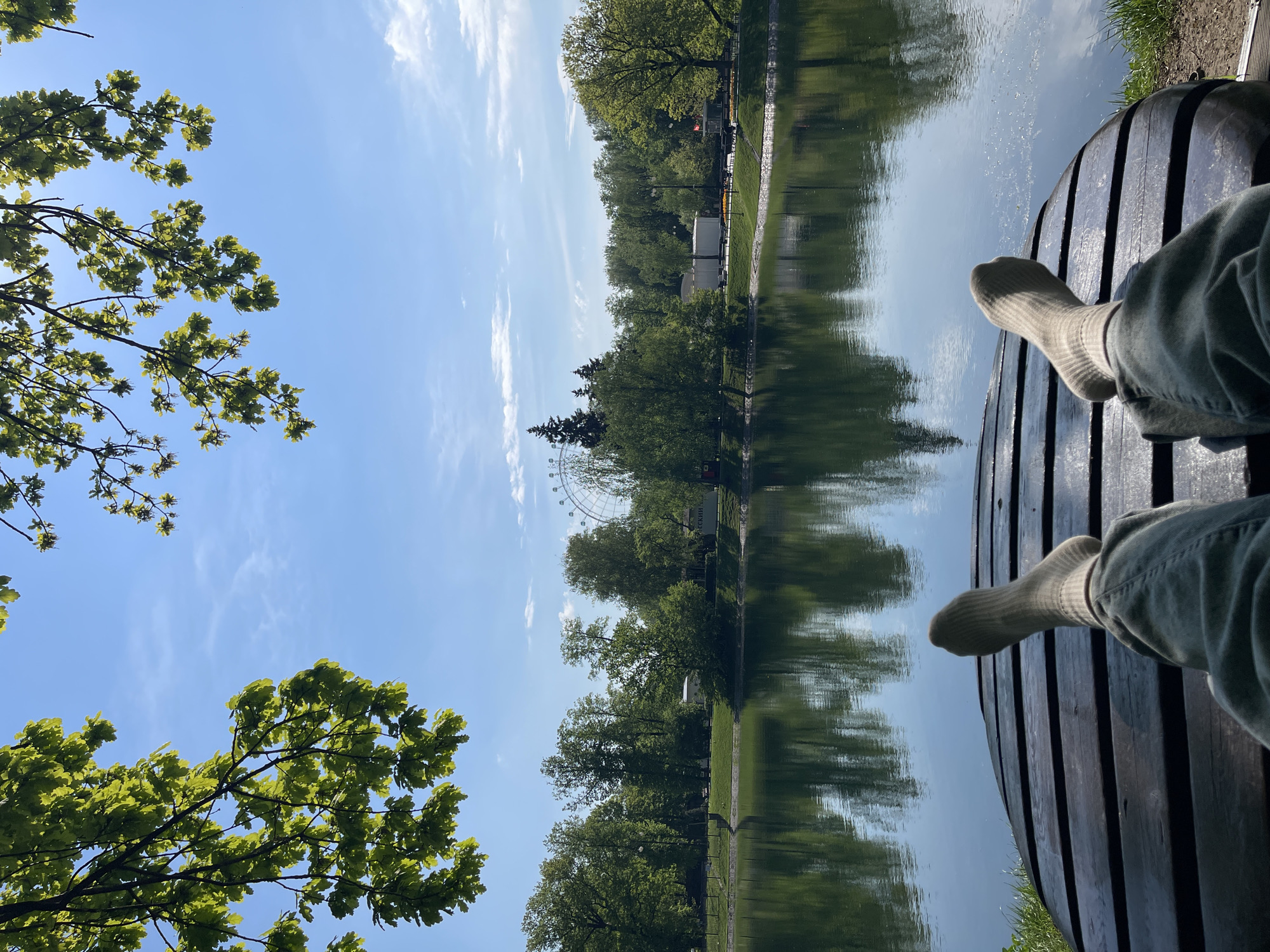 A vertical eye-level shot captures a person relaxing on a wooden bench beside a pond in an outdoor urban setting, potentially in Moscow, Russia. The individual, wearing socks and light-colored jeans, has their feet extended towards the water. The pond reflects the trees and sky, creating a mirror effect. Across the pond, trees line the edge, and a Ferris wheel is visible in the background. The sky is blue with scattered clouds. Green foliage from nearby trees fills the lower-left corner of the frame. The shot is taken in daylight, with clear visibility.