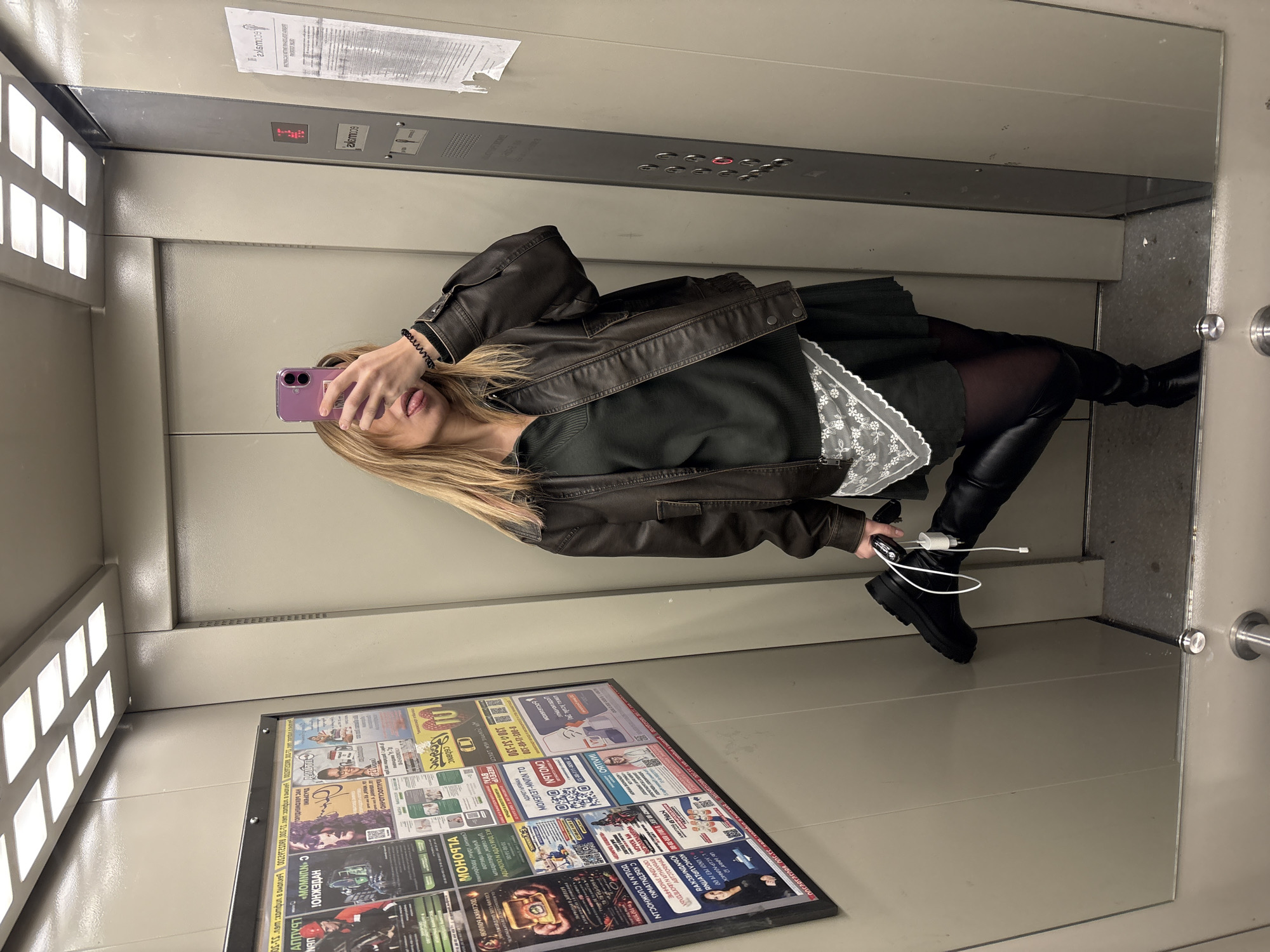 A woman is taking a selfie in a mirrored elevator. She is wearing a brown leather jacket, a dark green dress with a lace detail, black tights, and black, chunky boots. Her blonde hair is shoulder-length. She is holding a purple cell phone and sticking her tongue out. Her phone is connected to a charger. In the elevator, there is a display with multiple advertisements in Russian. The location is Ust-Ilimsk, Russia, based on the Cyrillic text in the advertisements. There is an elevator control panel above her. The scene is brightly lit by the elevator's lighting.