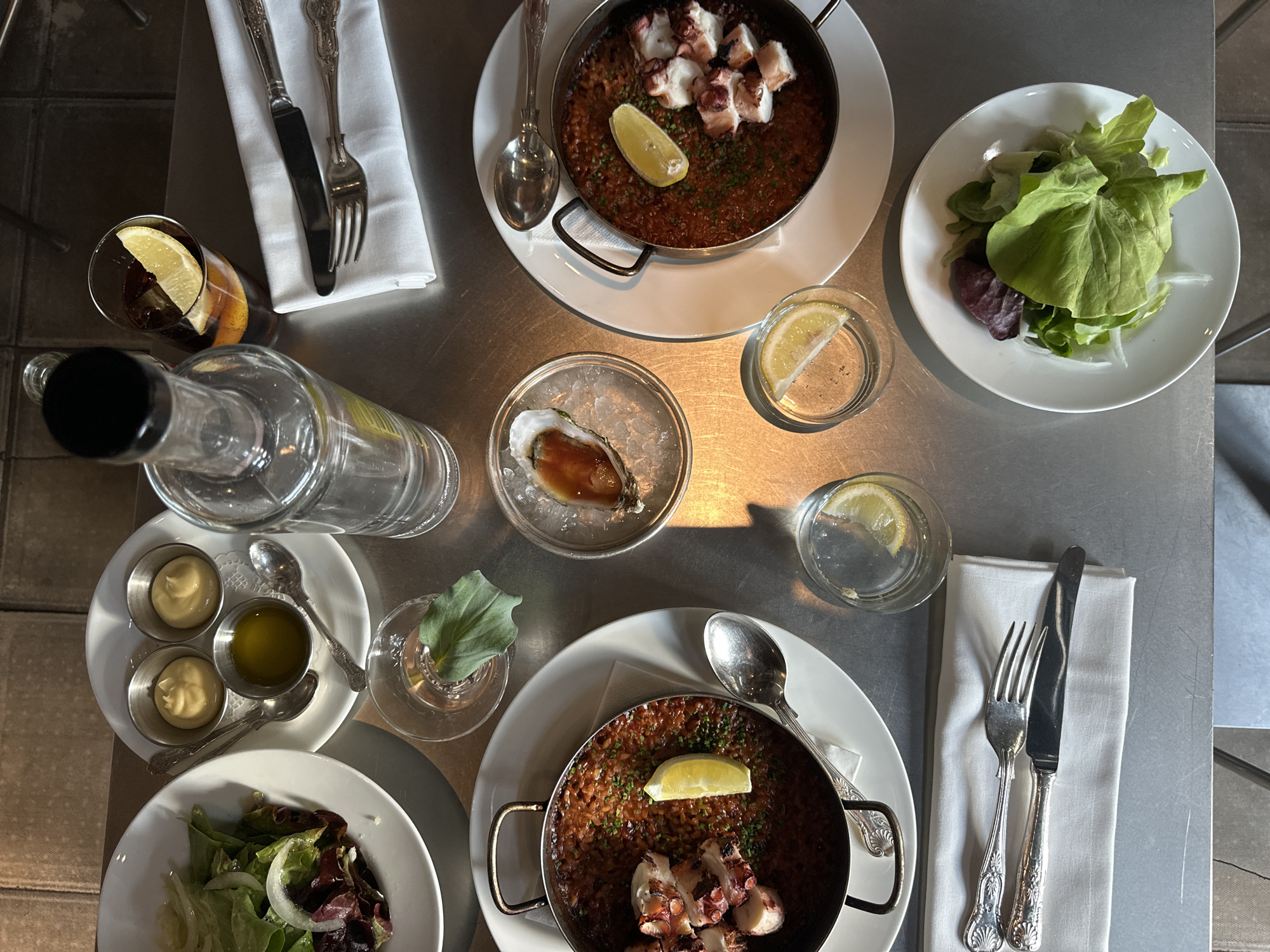 The image shows an overhead view of a restaurant table setting. The table is a brushed metal surface. There are two identical paella dishes containing rice and octopus, each garnished with a lemon wedge and served on a white plate with a spoon. There are also two white plates with green salads. One plate holds an oyster on ice. There are two glasses of water, each with a lemon wedge. There are small dishes of sauces and olive oil on a white plate with two small spoons. A bottle of clear liquid, possibly water, lies horizontally on the table. There are two sets of silverware wrapped in white napkins. One glass of dark liquid with a lemon garnish is visible. The location may be in the city of London, United Kingdom.
