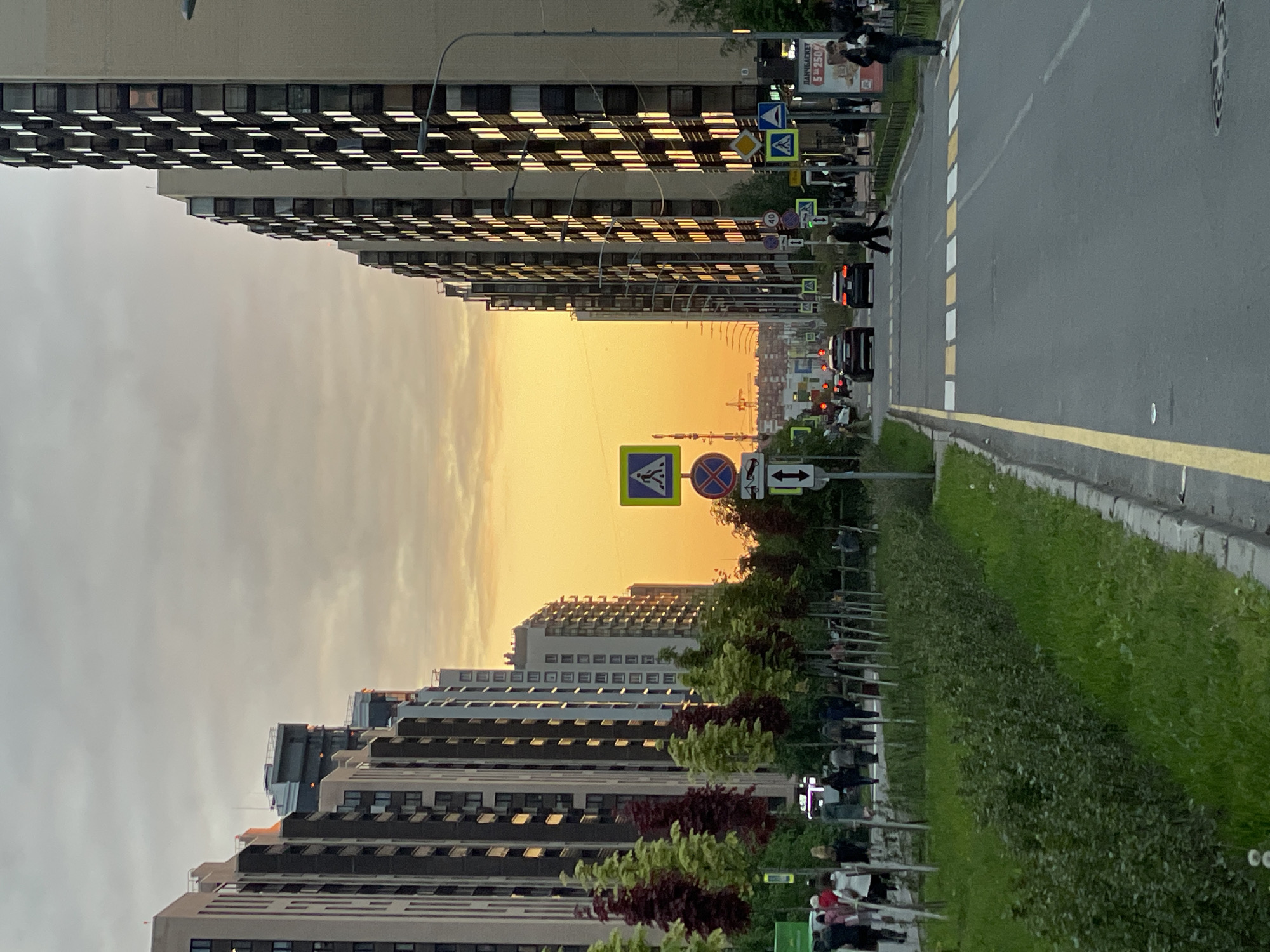 The image displays a city street viewed longitudinally towards a vibrant orange and yellow horizon, likely at sunset or sunrise. Tall, modern apartment buildings with numerous windows line both sides of the asphalt road, their facades and windows reflecting the warm glow from the sky.
The street features painted white and yellow lines, including a marked pedestrian crosswalk where one person is seen actively crossing. Several other pedestrians are scattered along the sidewalks and further down the street. A few cars are visible on the road, some with illuminated taillights.
A green median strip with grass, hedges, and small trees separates the road from the sidewalk. Numerous road signs are present, including those indicating a pedestrian crossing (yellow diamond, blue square), directional arrows, and a "no stopping/parking" symbol (blue circle with red diagonal line). A commercial sign in the upper right section of the image (when viewed upright) shows the text "ПАРКБАГЕТ" (Parkbaget) and "S > 250/".
The overall scene captures a typical urban environment during the golden hour, with the striking sky providing a focal point at the end of the long street.