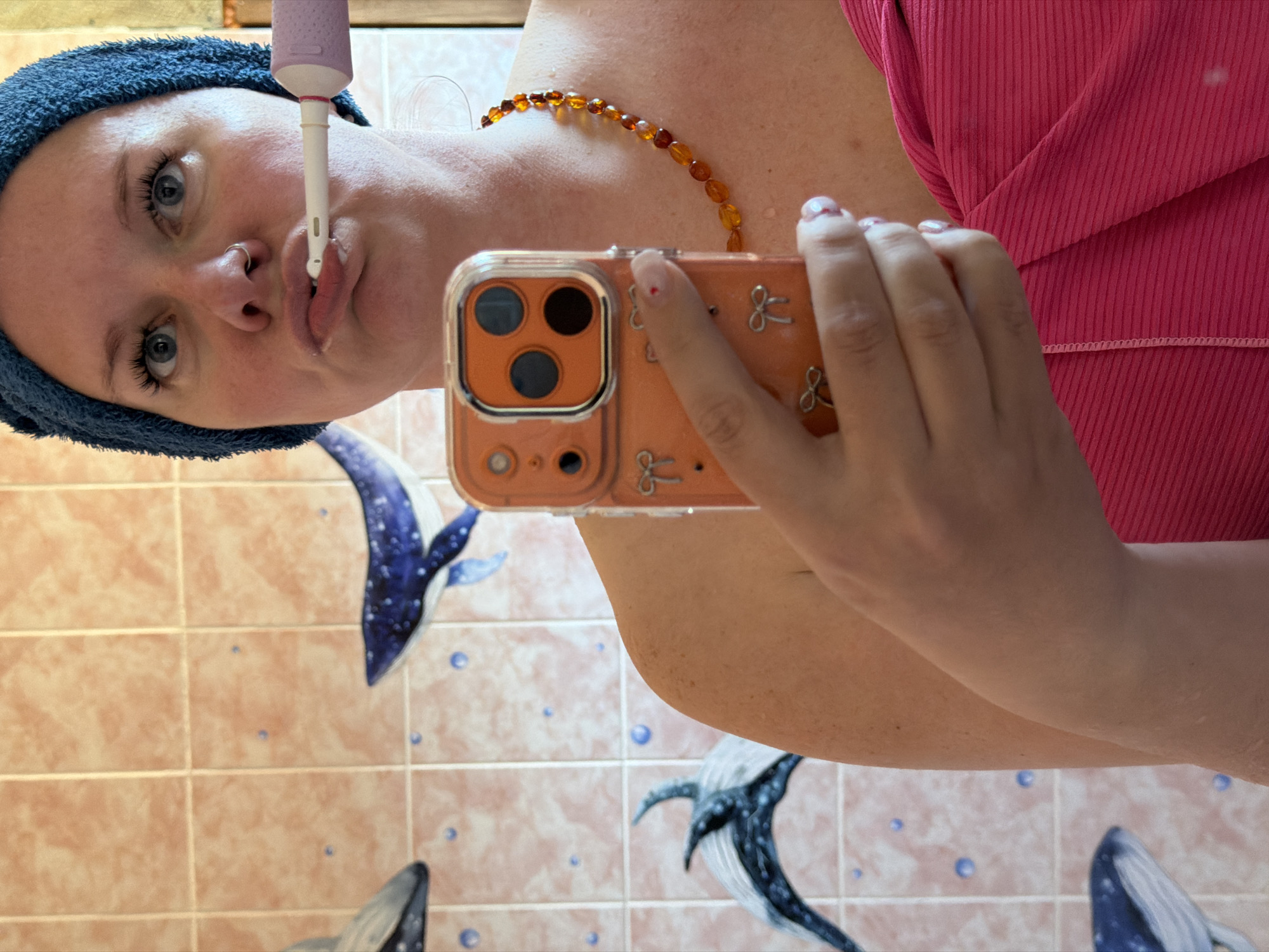 A selfie image depicts a woman holding an orange-cased iPhone in her right hand, partially obscuring her face. She is wearing a blue towel on her head and a brown beaded necklace. An electric toothbrush is positioned near her mouth. She is wearing a pink top. The background features light pink tiles decorated with blue whale motifs. The photo was taken in what appears to be a bathroom. There is no evidence to place this image in Chaikovsky, Russia.