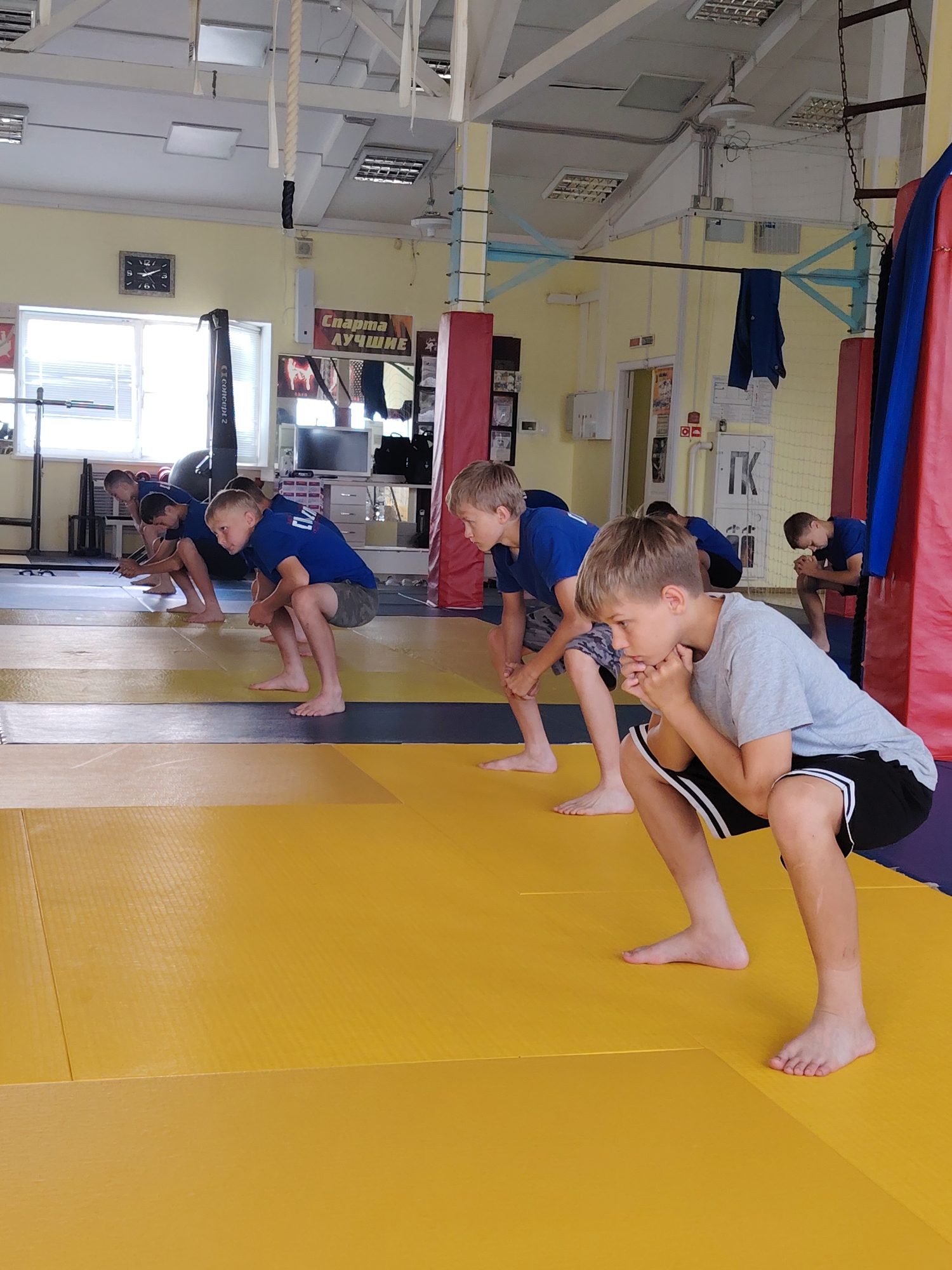 In a dojo located in Chaikovsky, Russia, a group of young boys is engaged in a physical training session on a yellow mat. The boys, dressed in blue t-shirts and patterned shorts or camouflage shorts, are performing a squatting exercise, with their bodies crouched low and hands often resting near their faces. One boy in the foreground is wearing a grey t-shirt and black shorts with white stripes. The environment features a high ceiling with exposed beams and lighting fixtures, a clock on the wall, a window, and sports equipment such as a rowing machine, punching bags, and a climbing rope. Signs with Russian text are visible on the walls, suggesting the location is a sports facility. The overall scene depicts a disciplined training environment.