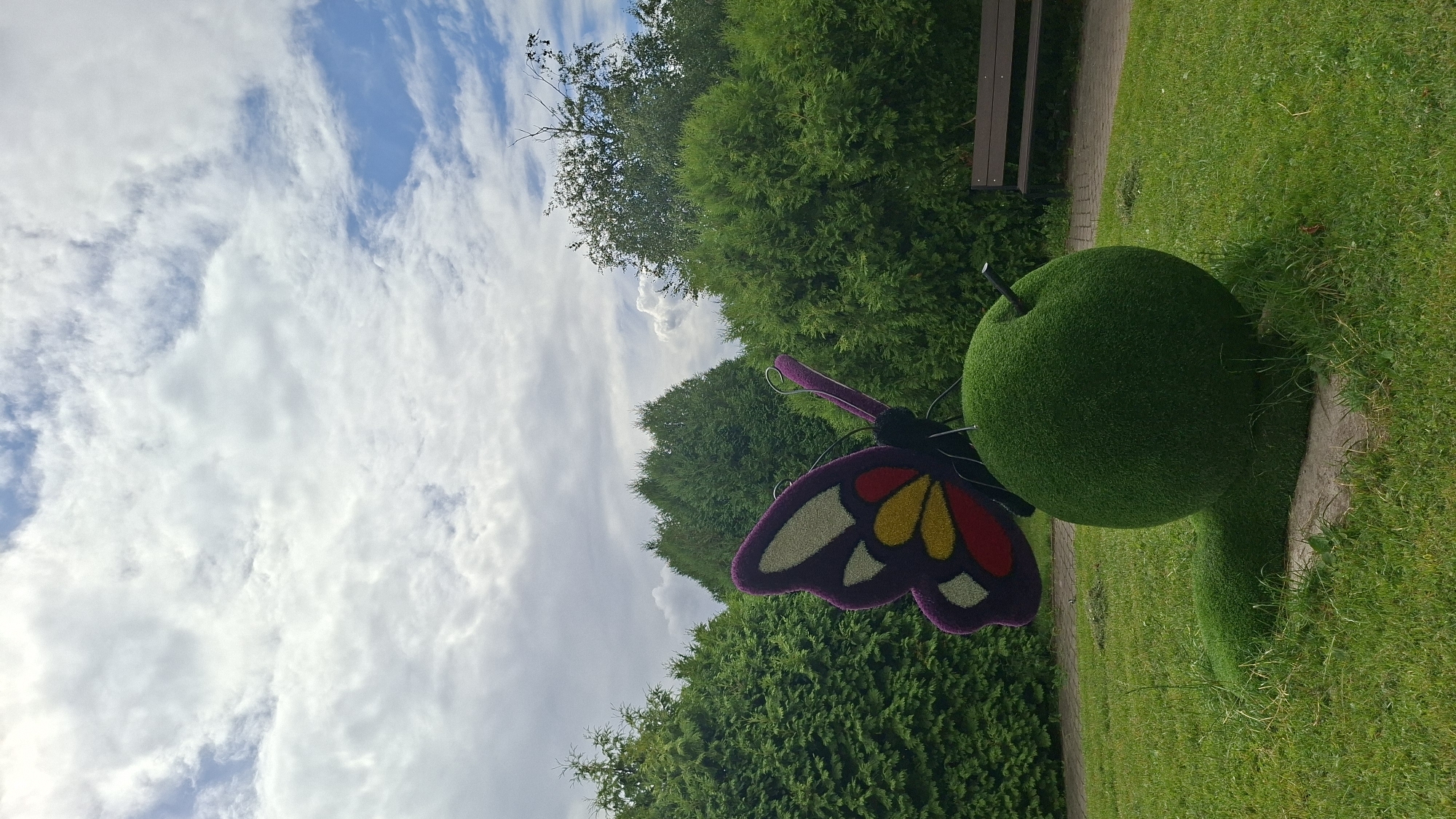 This is a medium-angle, ground-level, outdoor, daytime, full-shot photograph of a decorative topiary sculpture of a caterpillar with a butterfly on its back in a park. The sculpture is made of green artificial grass and is situated on a paved walkway, with its body extending into the surrounding lawn. The caterpillar's body is rounded and segmented, while its head is a large, smooth sphere. Perched on top of the sphere is a vibrantly colored butterfly sculpture, featuring purple, yellow, and red wing patterns. The background consists of dense, lush green trees and shrubs, suggesting a well-maintained park or garden. In the upper portion of the image, a cloudy sky with patches of blue is visible. To the right of the sculpture, a wooden park bench is partially visible, also situated near a paved walkway. The overall impression is of a whimsical, artistic garden feature. The photograph was taken in the city of Myadzyel, Belarus.